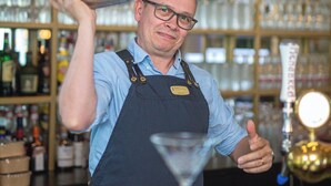 Un bar con estantes llenos de botellas y vasos, un barman preparando una bebida, y una barra.