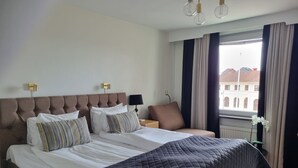 Habitación de hotel con una cama grande, una mesita de noche, una lámpara y vistas a edificios a través de la ventana.