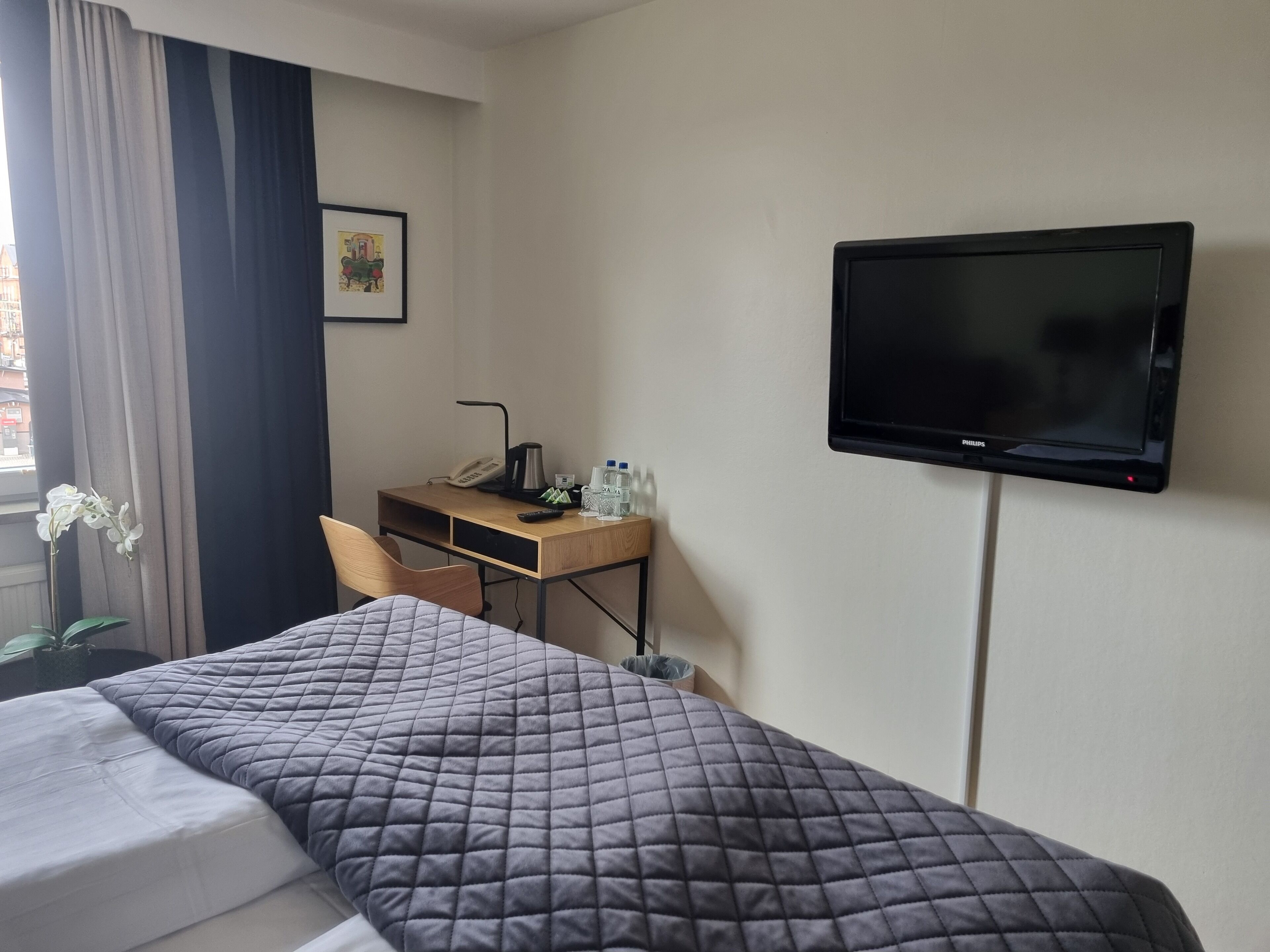 Quarto de hotel com cama, escrivaninha com telefone, televisão e janela com cortinas.