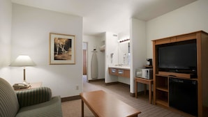Habitación de hotel con sofá a rayas verdes, una mesa de madera, un televisor, microondas, un refrigerador pequeño, tocador con espejo y un cuadro en la pared.