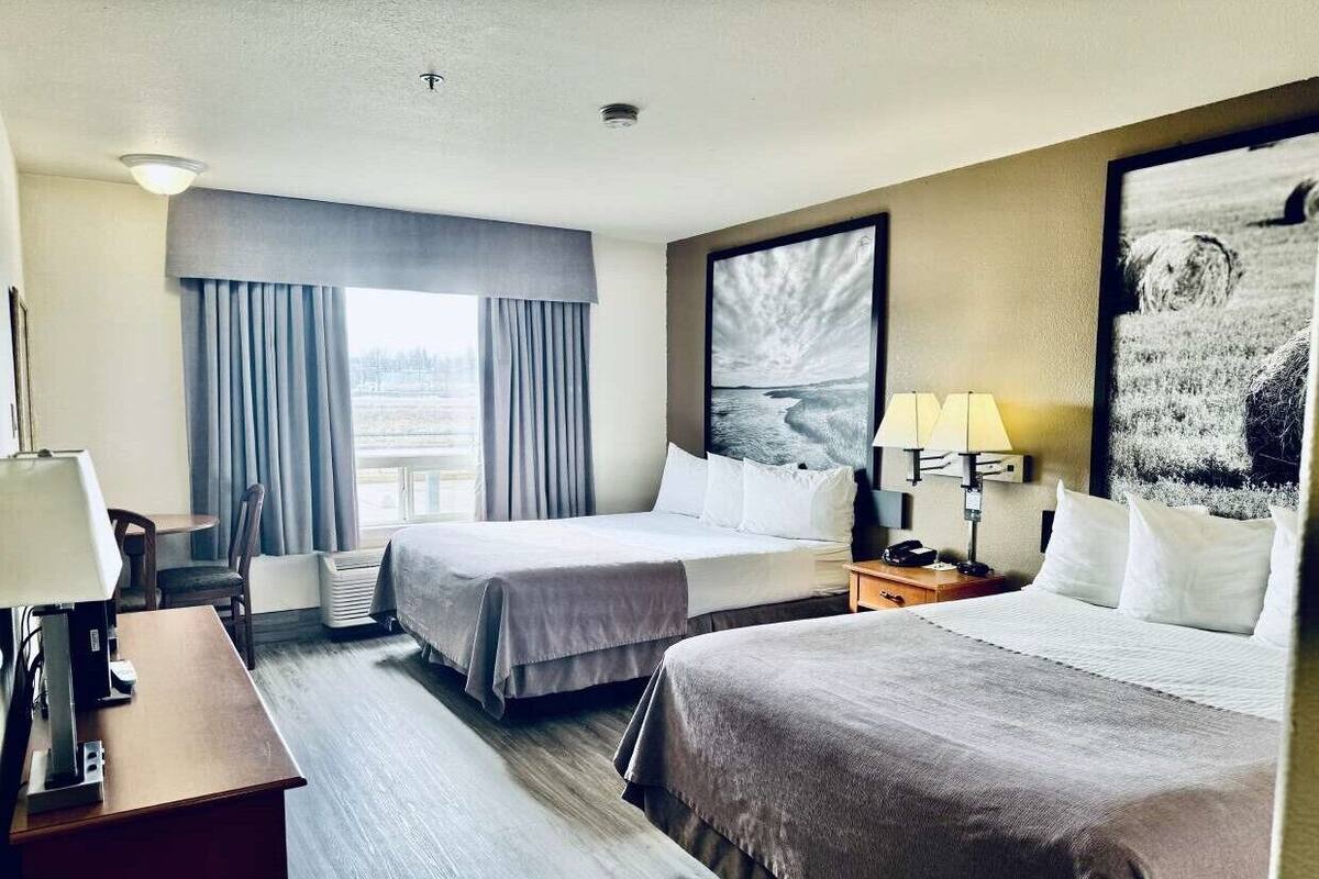 Habitació d'hotel amb dos llits, un escriptori, una cadira, una làmpada i una finestra gran amb cortines.