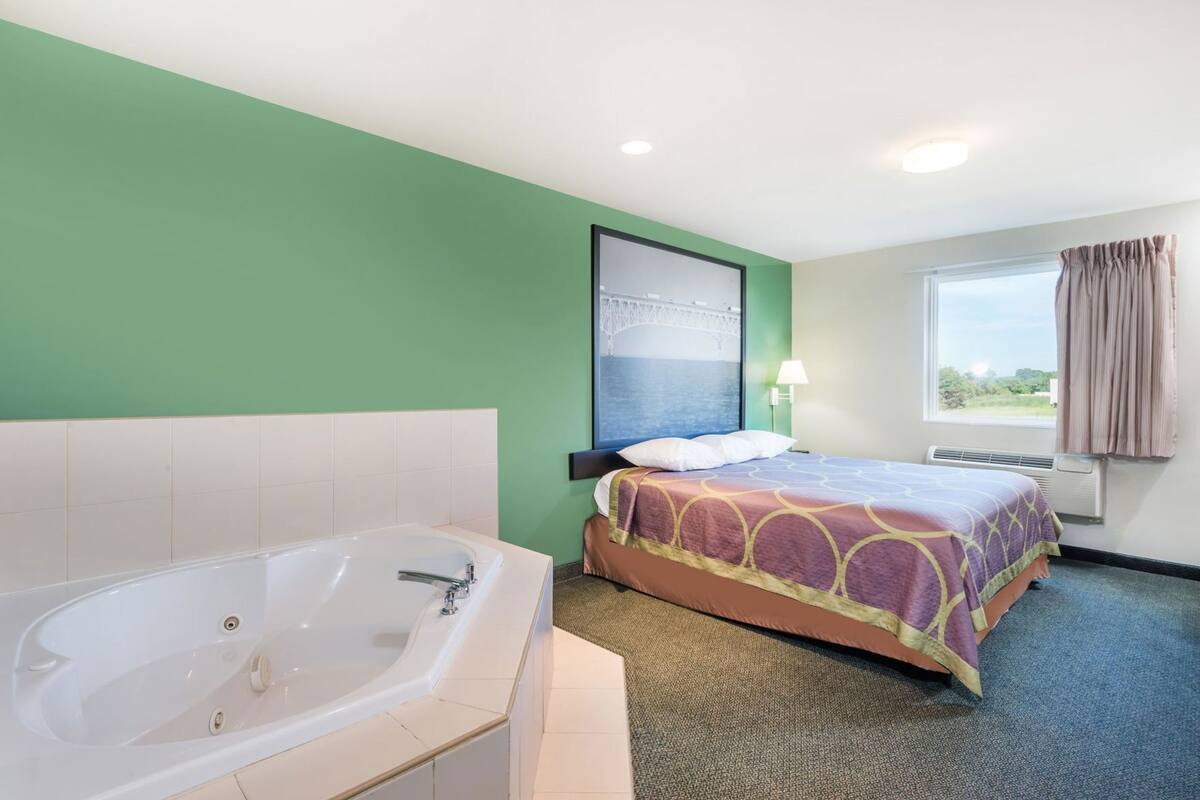 A hotel room with a large bathtub, a bed with a patterned bedspread, a nightstand with a lamp, and a window with curtains.
