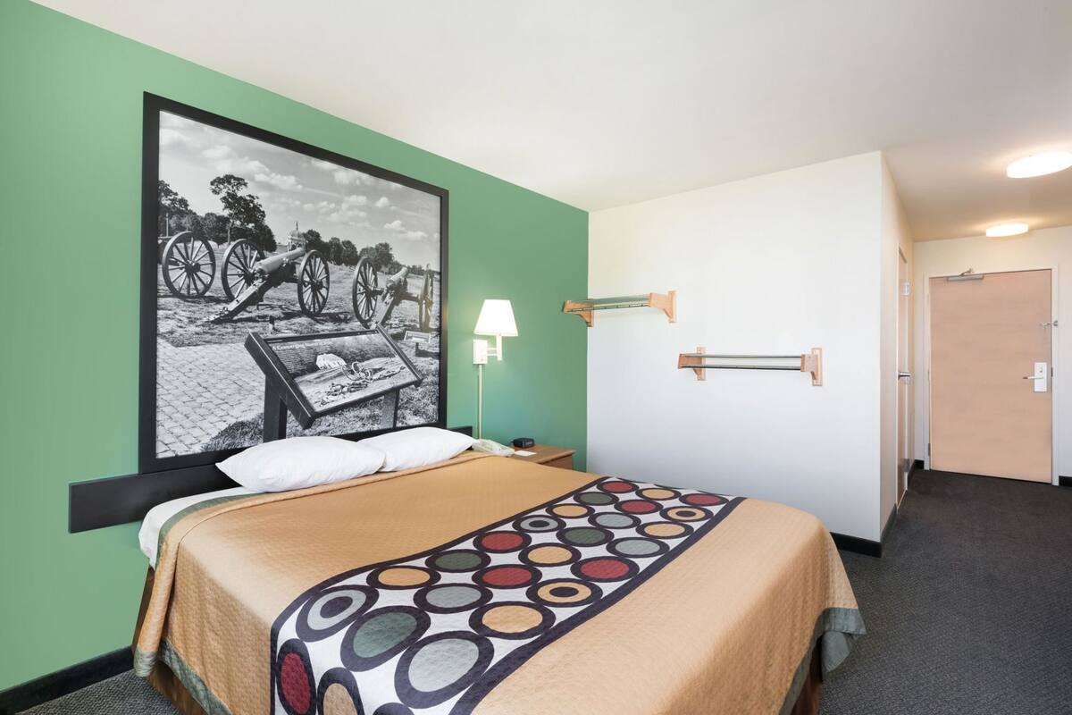 A hotel room with a bed, a wall-mounted lamp, a door, and a framed historical photo.