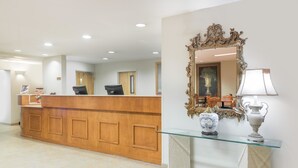 A reception area with a wooden reception desk, a glass-top console table with a lamp, and a large ornate mirror on the wall. - Super 8 by Wyndham Hagerstown/Halfway Area (Hagerstown)