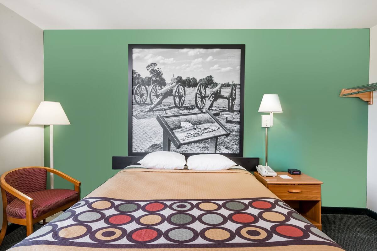 A hotel room with a bed, two bedside lamps, a chair, a wooden nightstand, and a large framed picture on the wall.