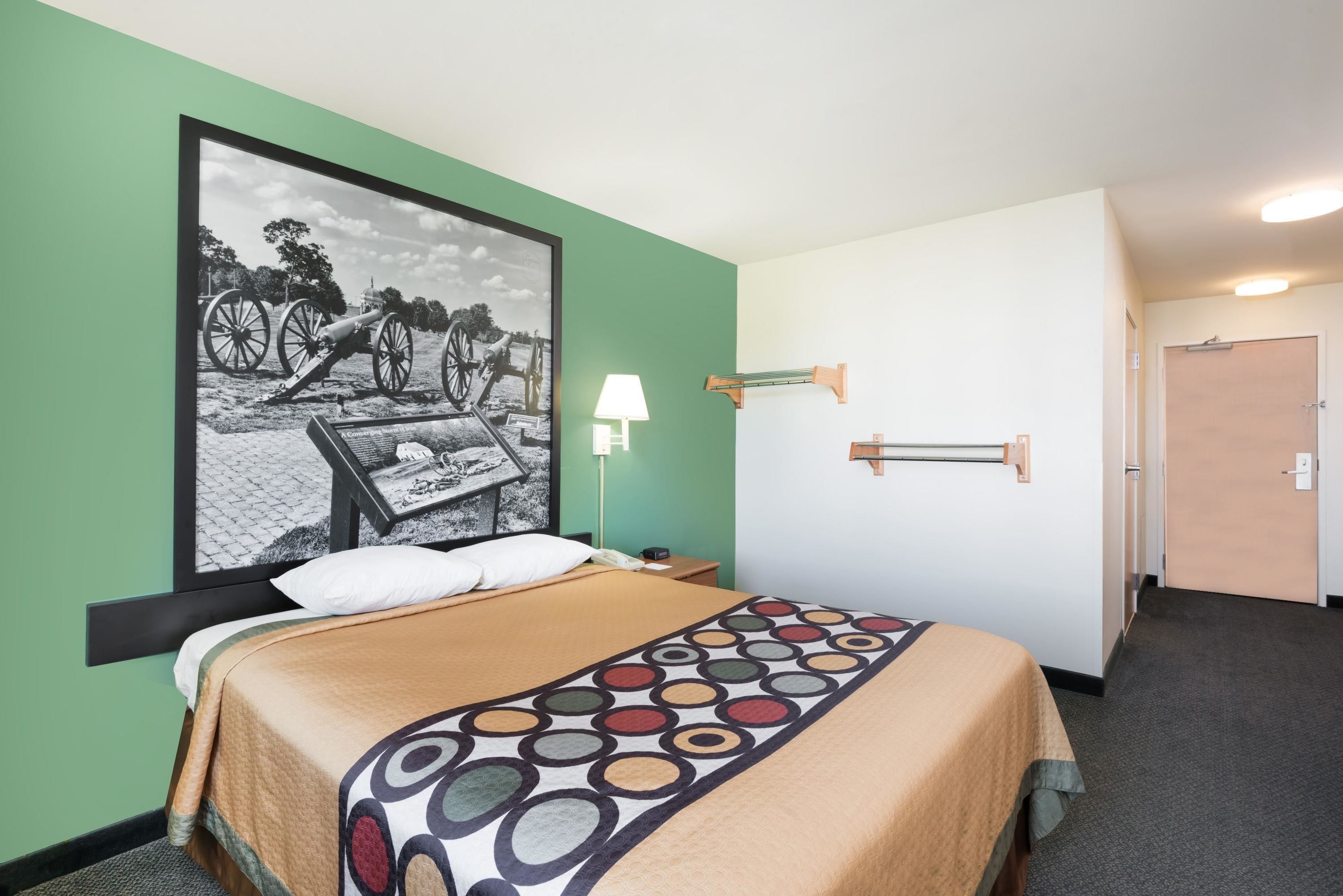 A hotel room with a bed, a nightstand, a lamp, a wall-mounted shelf, and a door.