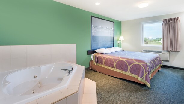 A hotel room with a large bathtub, a bed with a patterned bedspread, a window with curtains, and a wall-mounted picture. - Super 8 by Wyndham Hagerstown/Halfway Area (Hagerstown)