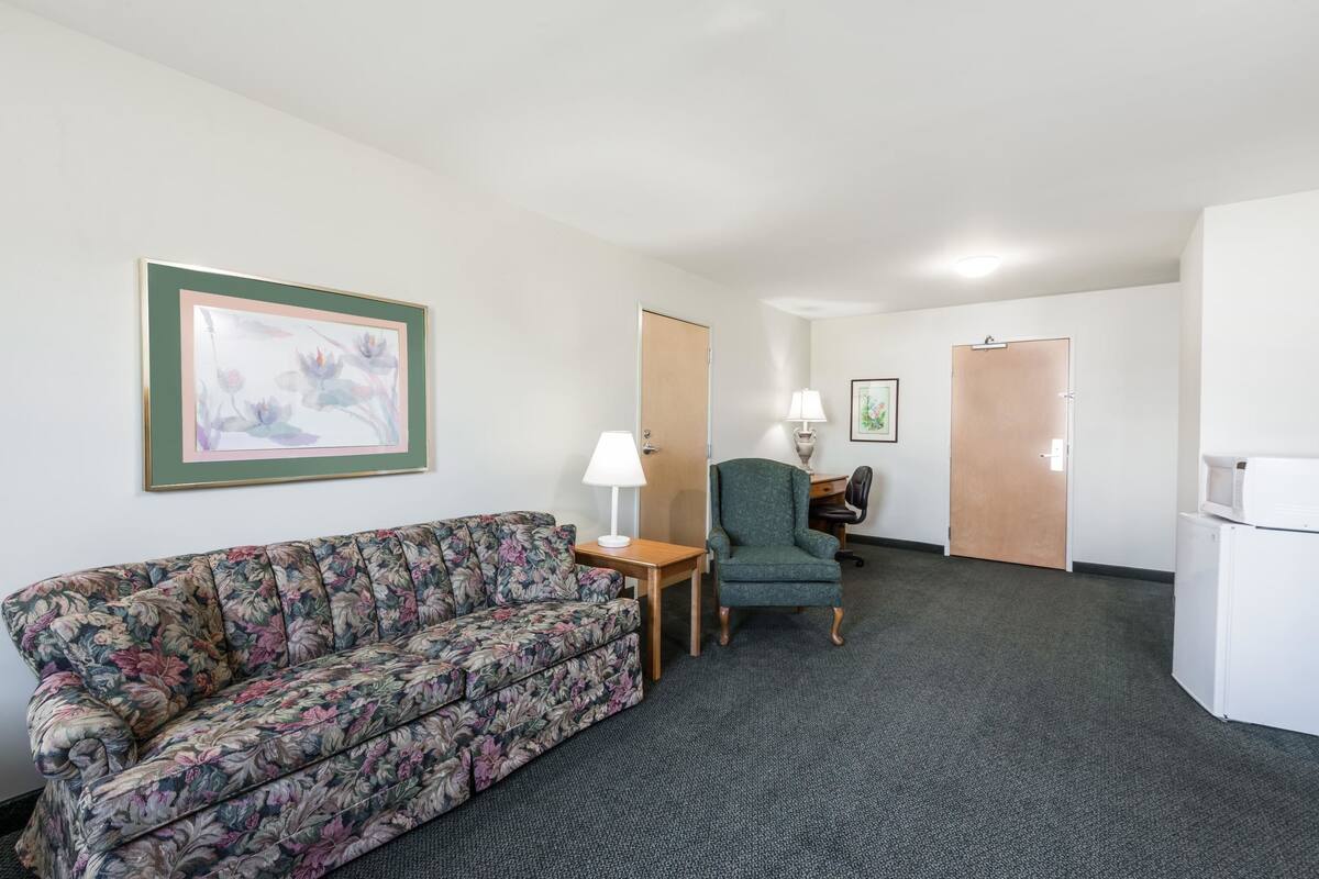 A hotel room with a floral-patterned sofa, a green armchair, a small table with a lamp, and a microwave on a cabinet.