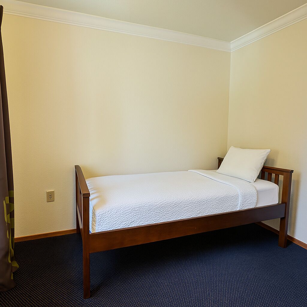 Una cama individual con estructura de madera y ropa de cama blanca en una habitación con paredes color beige y alfombra oscura.