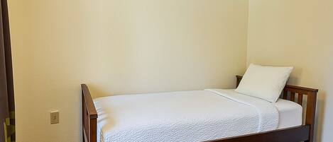 Una cama individual con estructura de madera y ropa de cama blanca en una habitación con paredes color beige y alfombra oscura.