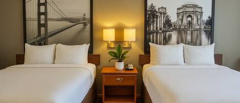 A hotel room with two beds, a nightstand with a lamp and a plant, and two framed pictures on the wall.