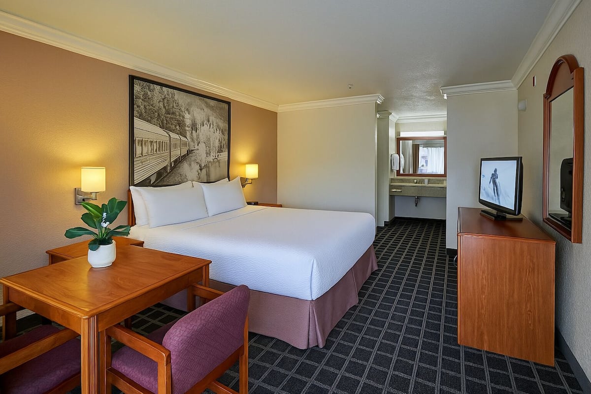 A hotel room with a bed, bedside table, chair, TV, and a painting on the wall.