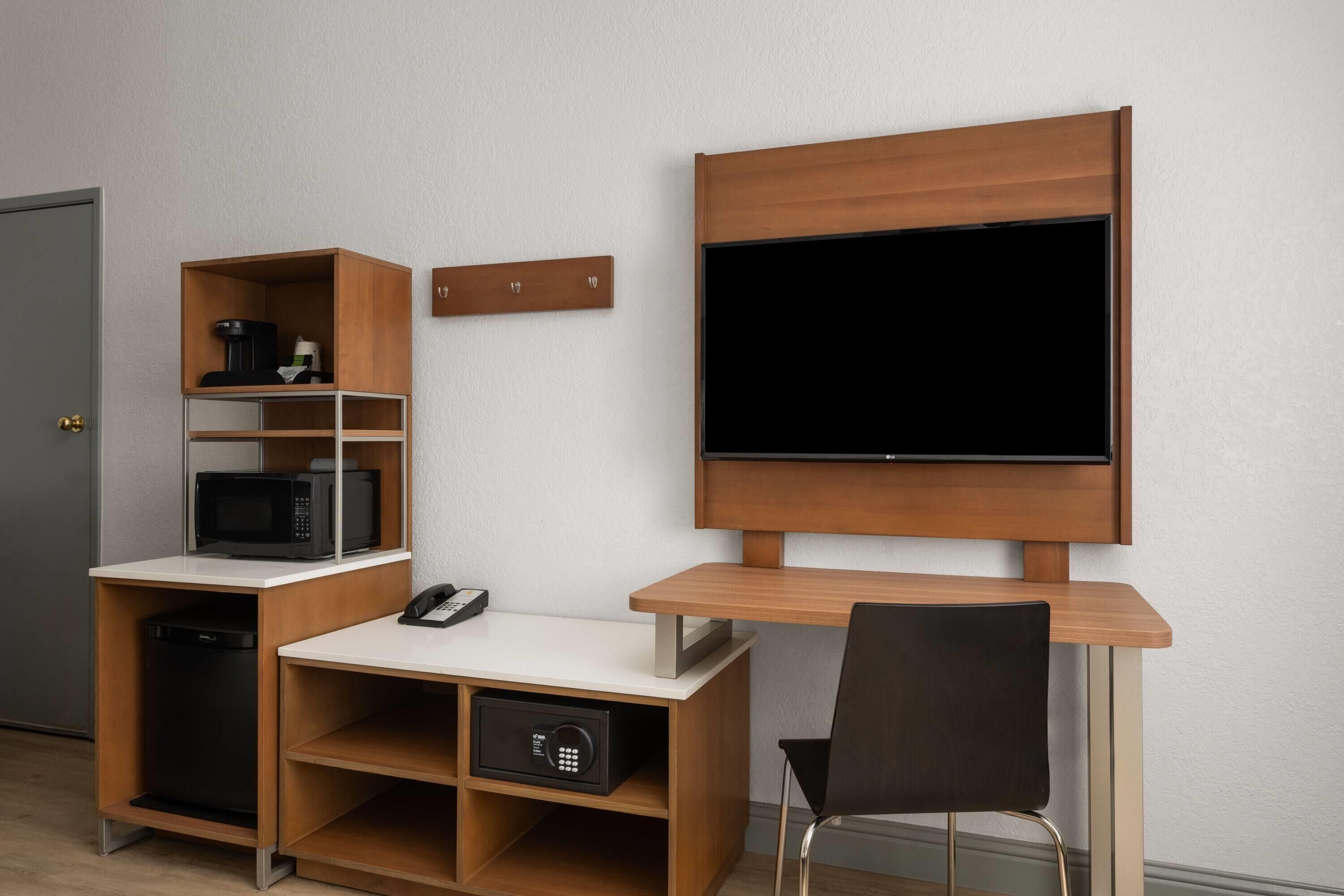 A compact room with a wooden desk, a mounted flat-screen TV, a microwave, and a coffee maker.