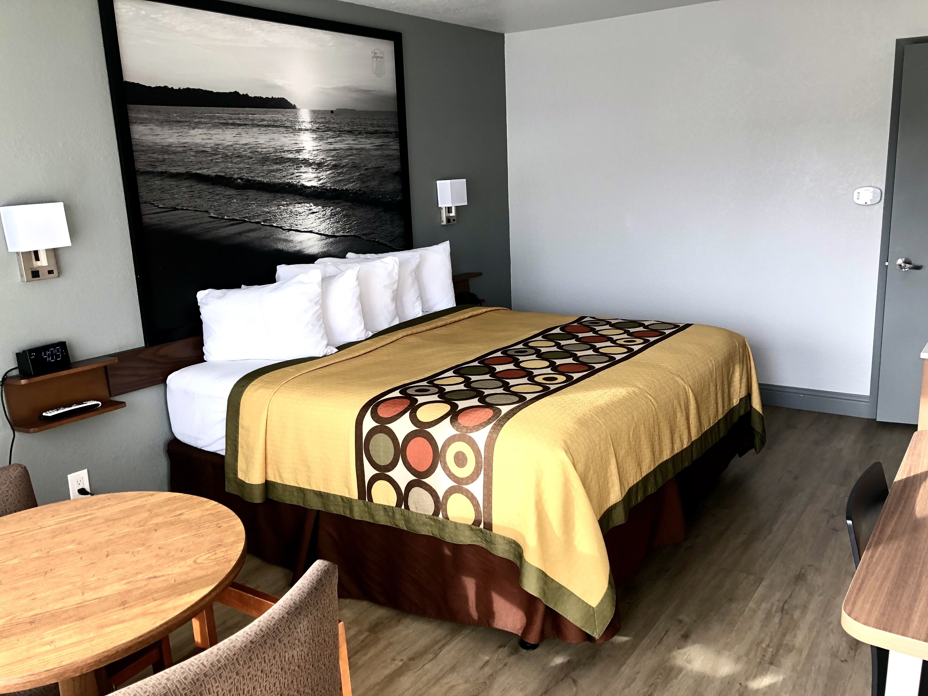 A hotel room with a bed, a desk, a chair, a round table, and a large painting of a beach scene.
