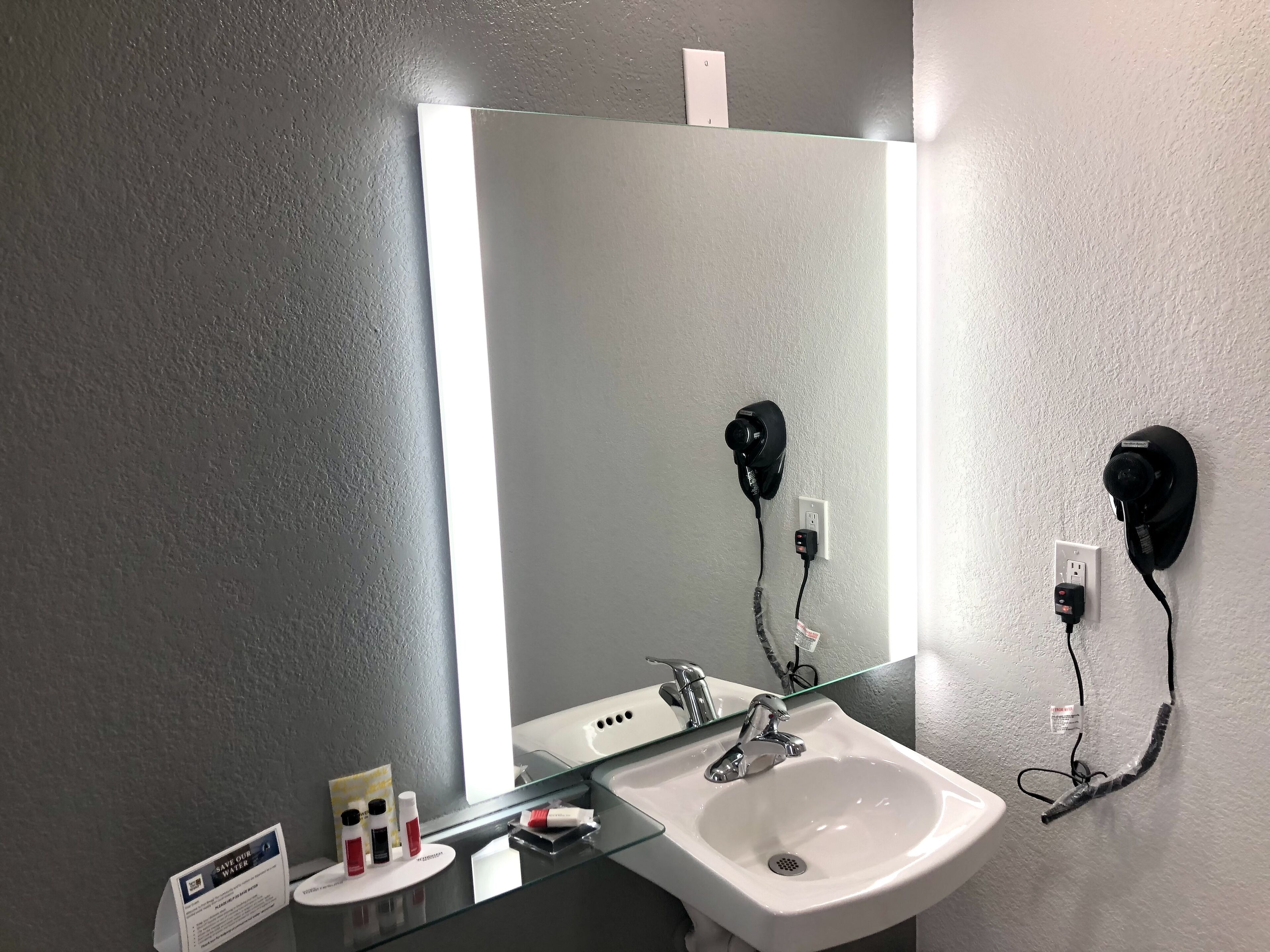 Een badkamer met een witte wastafel, een spiegel en twee haardrogers in het stopcontact. Er is een glazen plank met toiletartikelen en een bordje over waterbesparing.