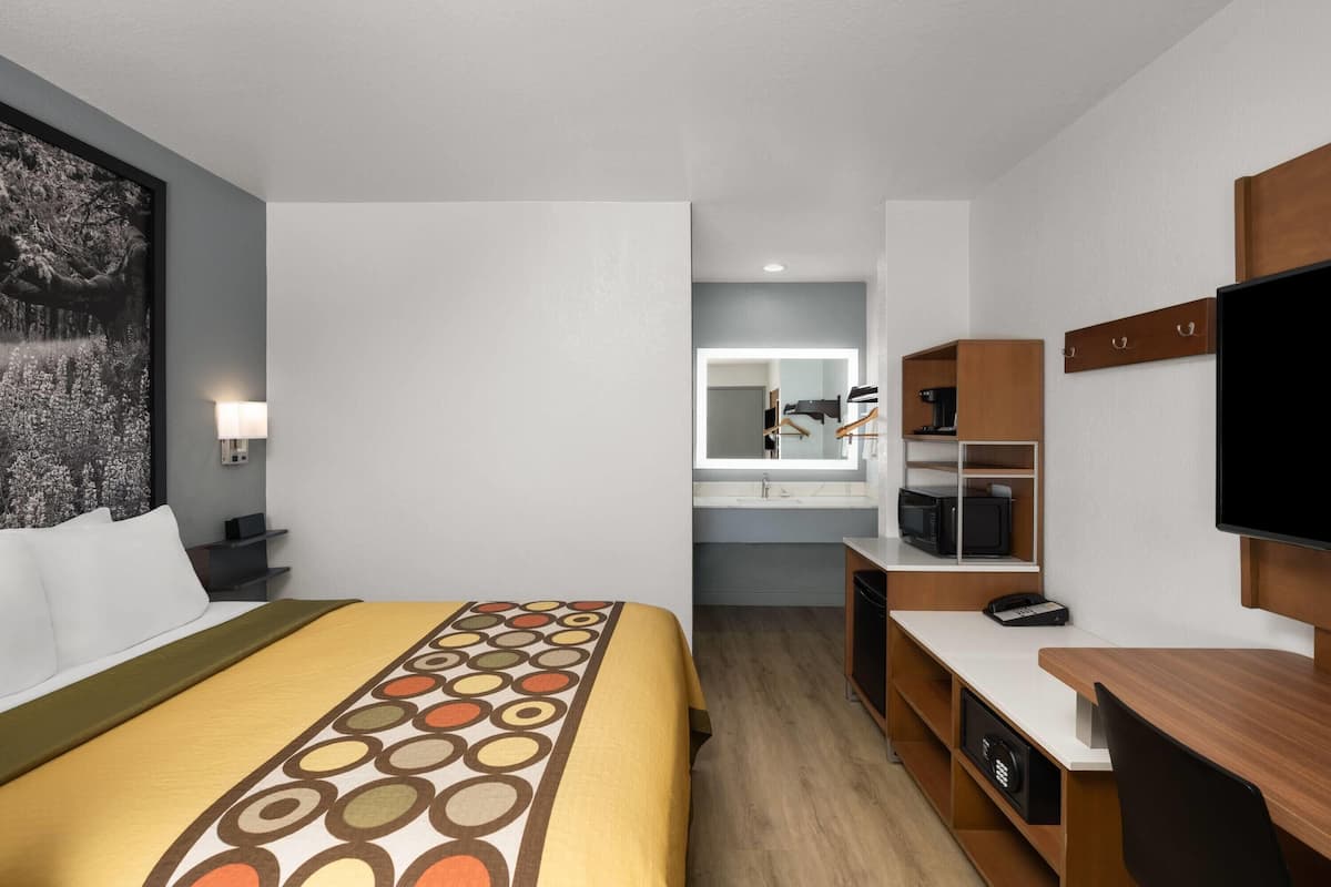 Ein Hotelzimmer mit Bett, Schreibtisch, Fernseher, Spiegel und einem Badezimmer mit Waschbecken und Mikrowelle.