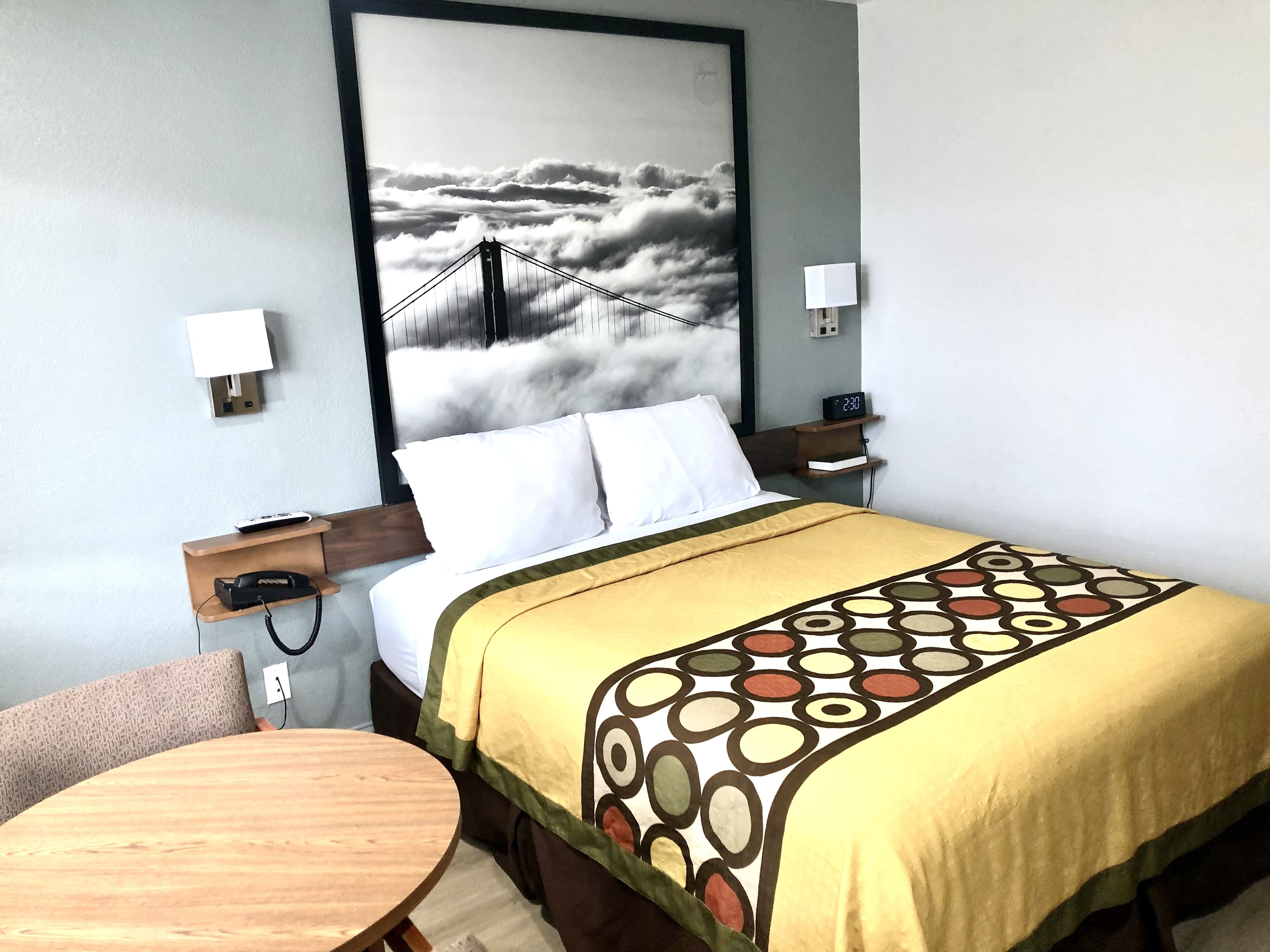 A hotel room with a bed, a round wooden table, a wall-mounted lamp, a digital clock, and a large framed picture of a bridge over clouds.