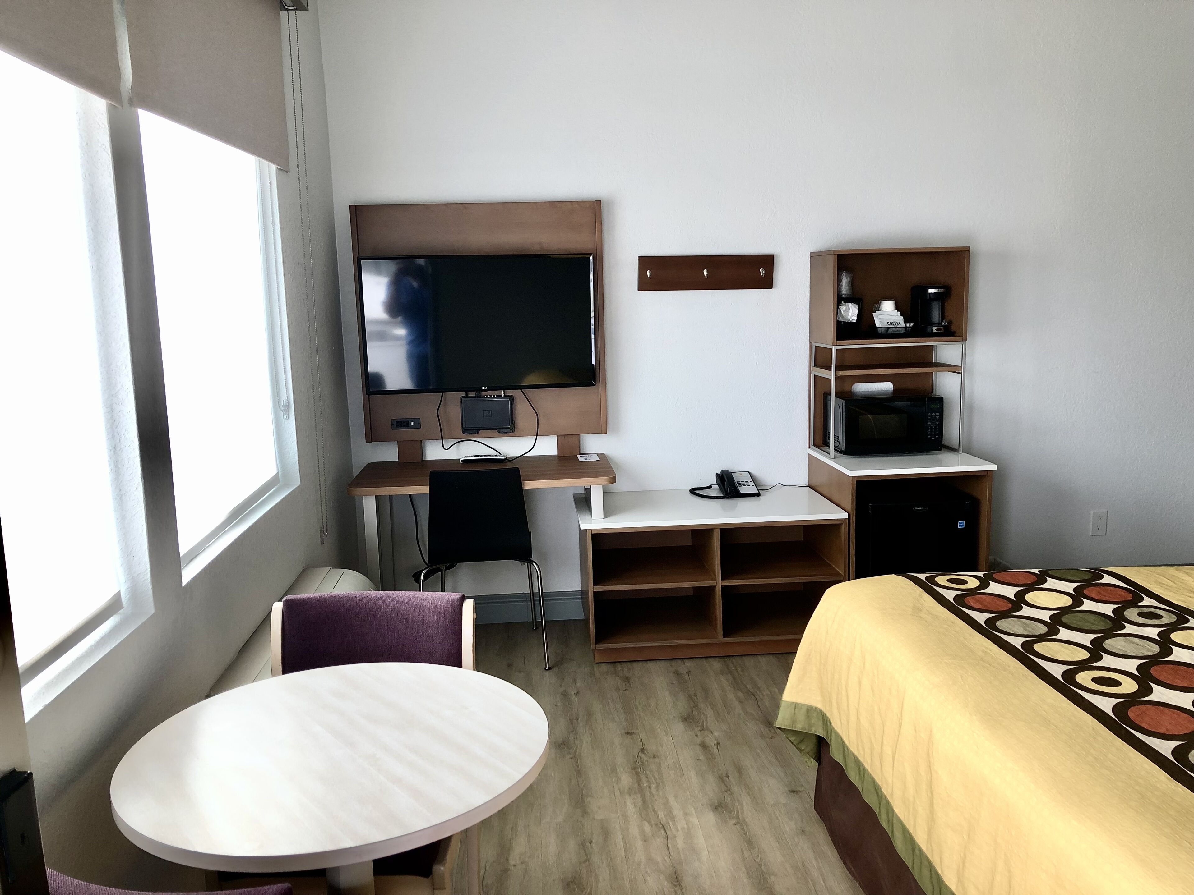 A hotel room with a bed, a desk with a TV, a chair, a small table, and a microwave.