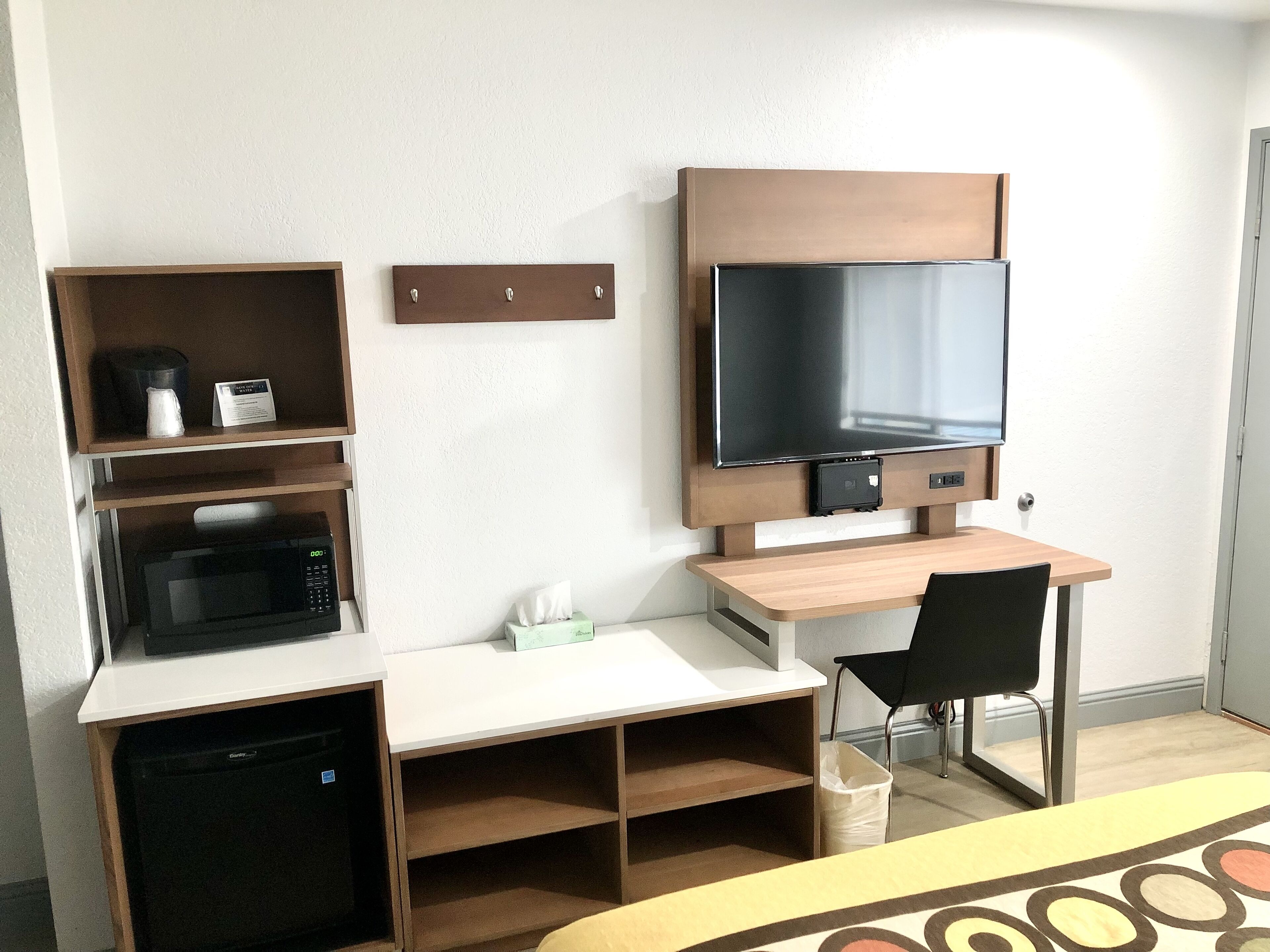A compact hotel room with a bed, a desk with a chair, a television, a microwave, and a small storage unit.