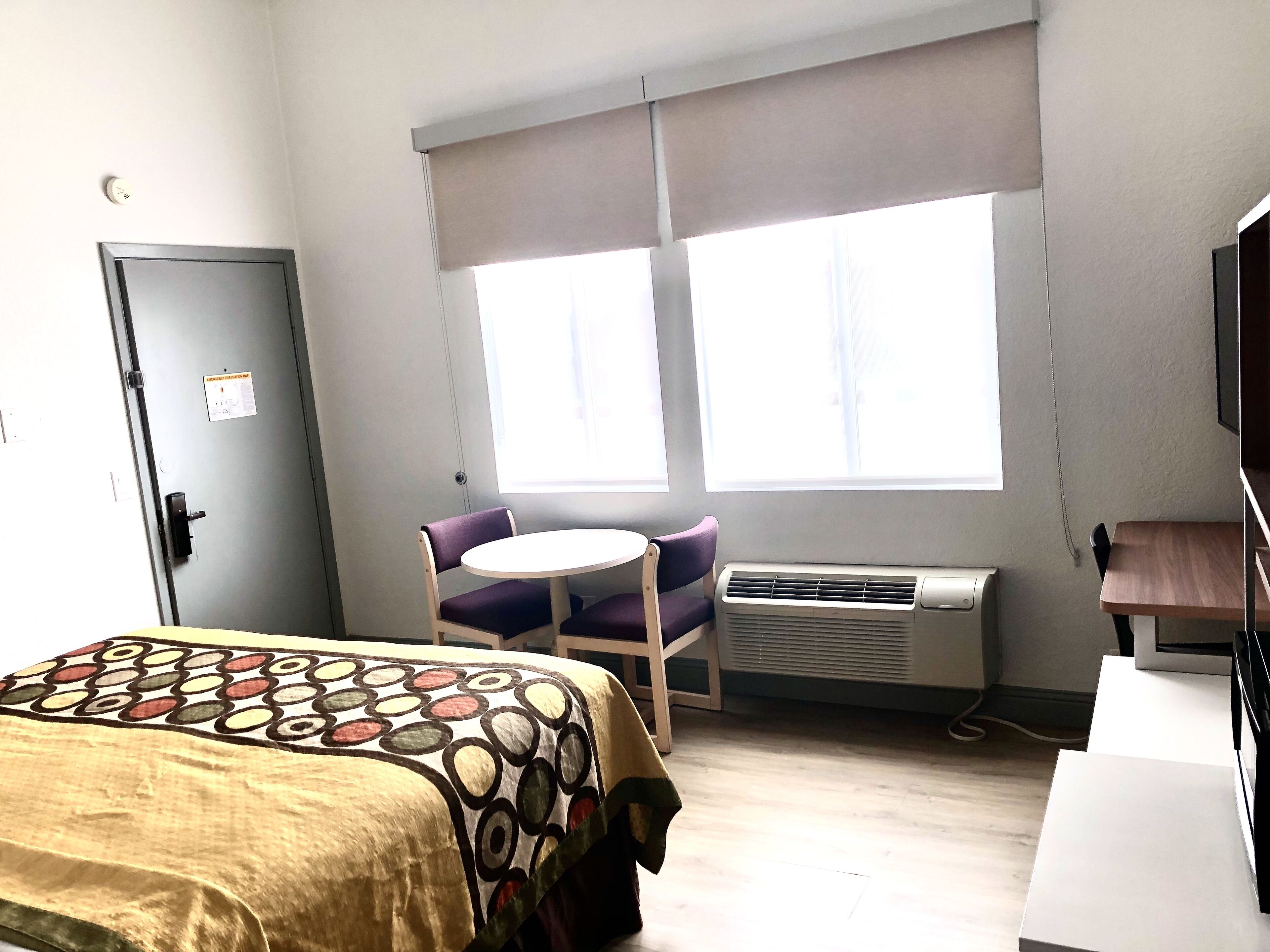 A hotel room with a bed, a desk, a chair, an air conditioner, and a window with blinds.