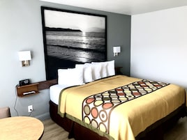 Habitación de hotel con cama, un reloj de pared, una mesa redonda y un cuadro grande con una playa.