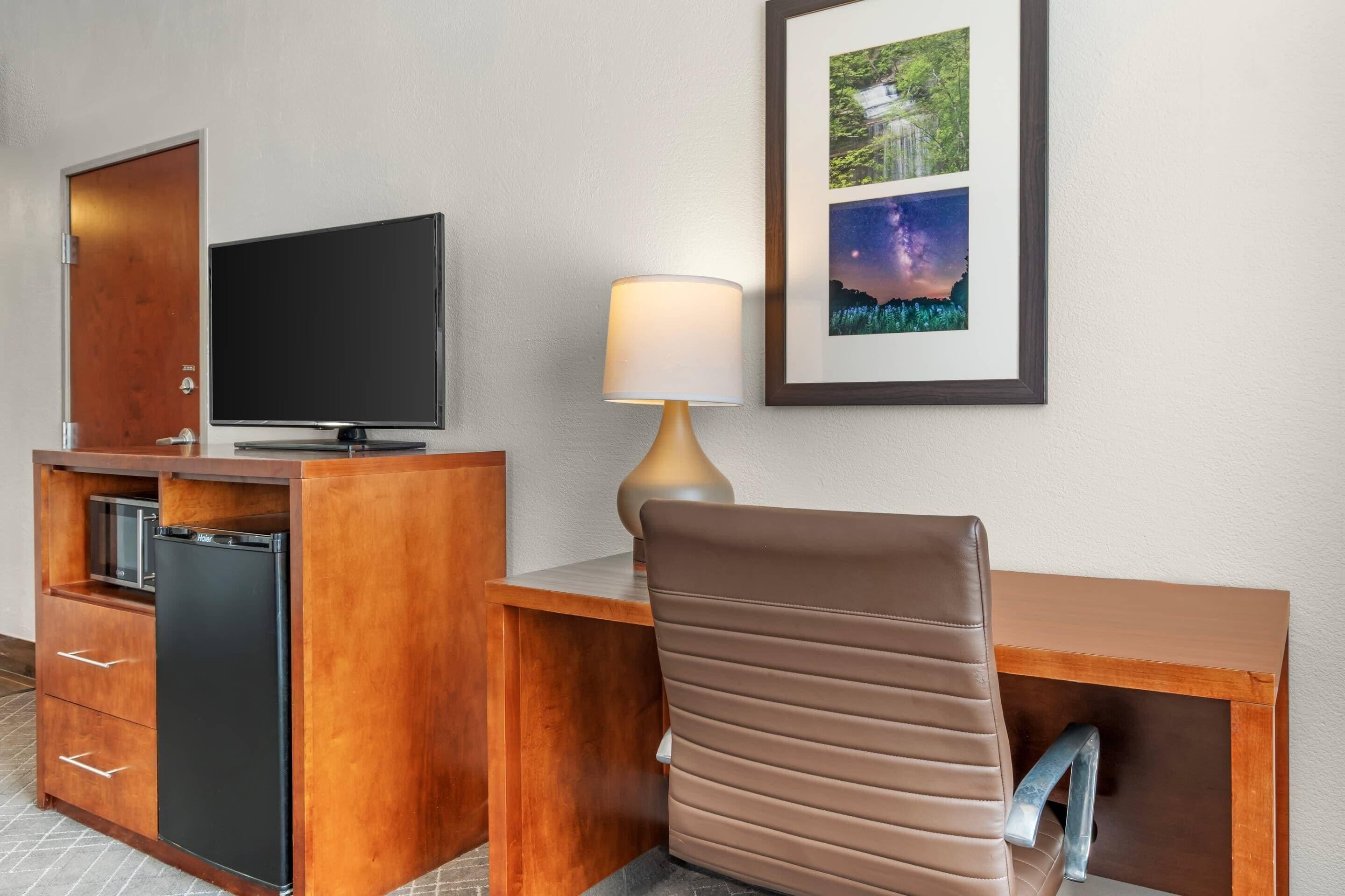 A hotel room with a wooden desk, a television, a lamp, a chair, a minibar, and a framed picture on the wall.
