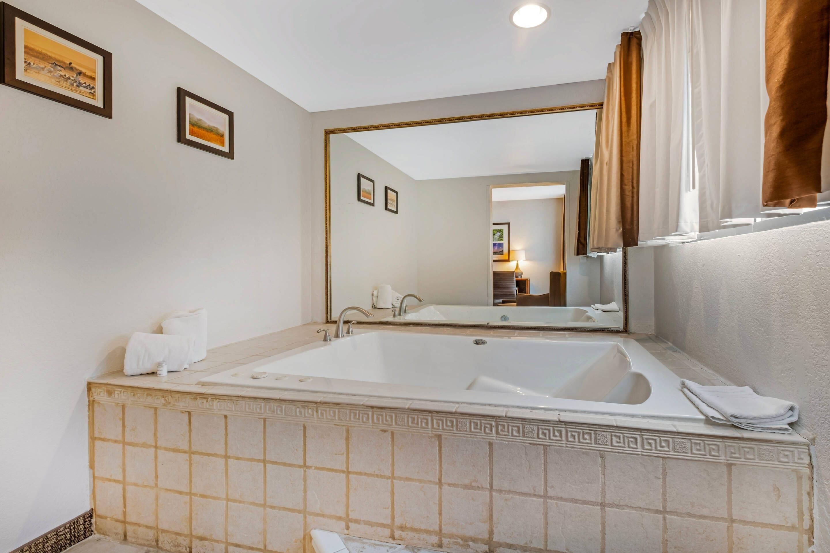 A bathroom with a large bathtub, a mirror, and a window with curtains.