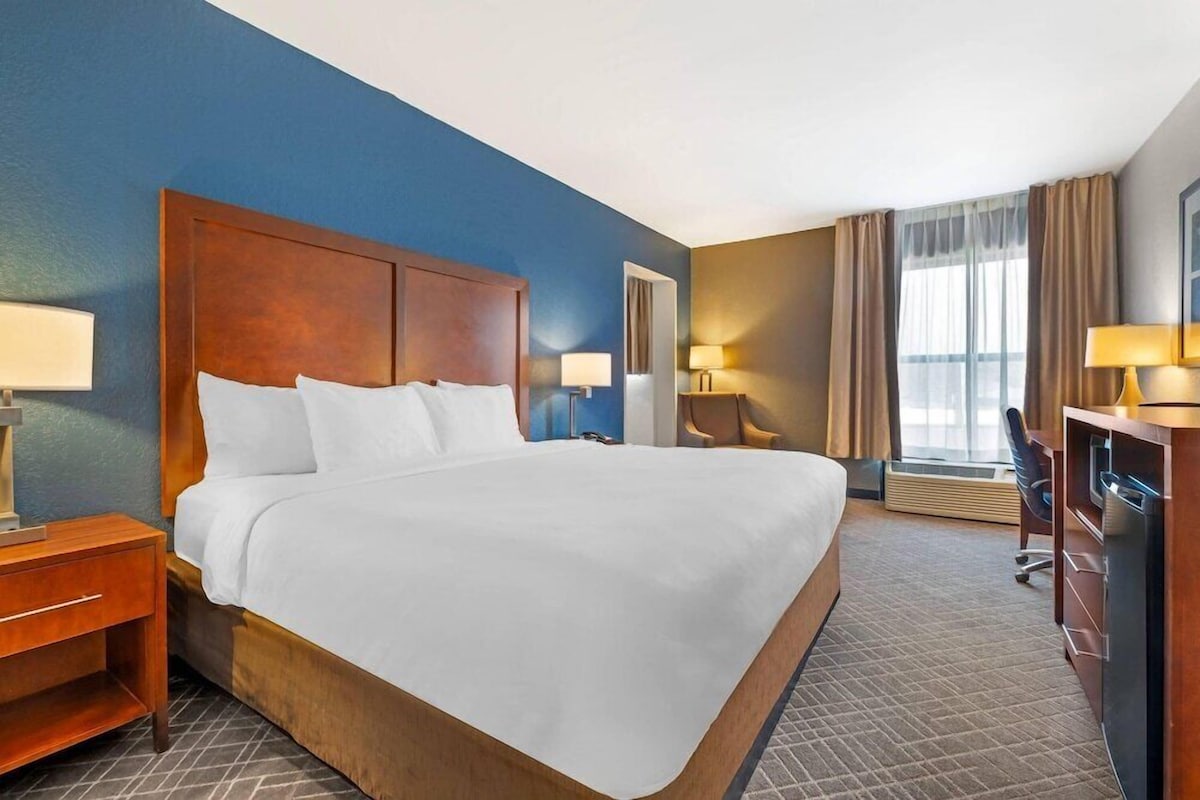 Habitación de hotel con una cama grande, dos mesitas de noche con lámparas, un escritorio con silla y un ventanal con cortinas.