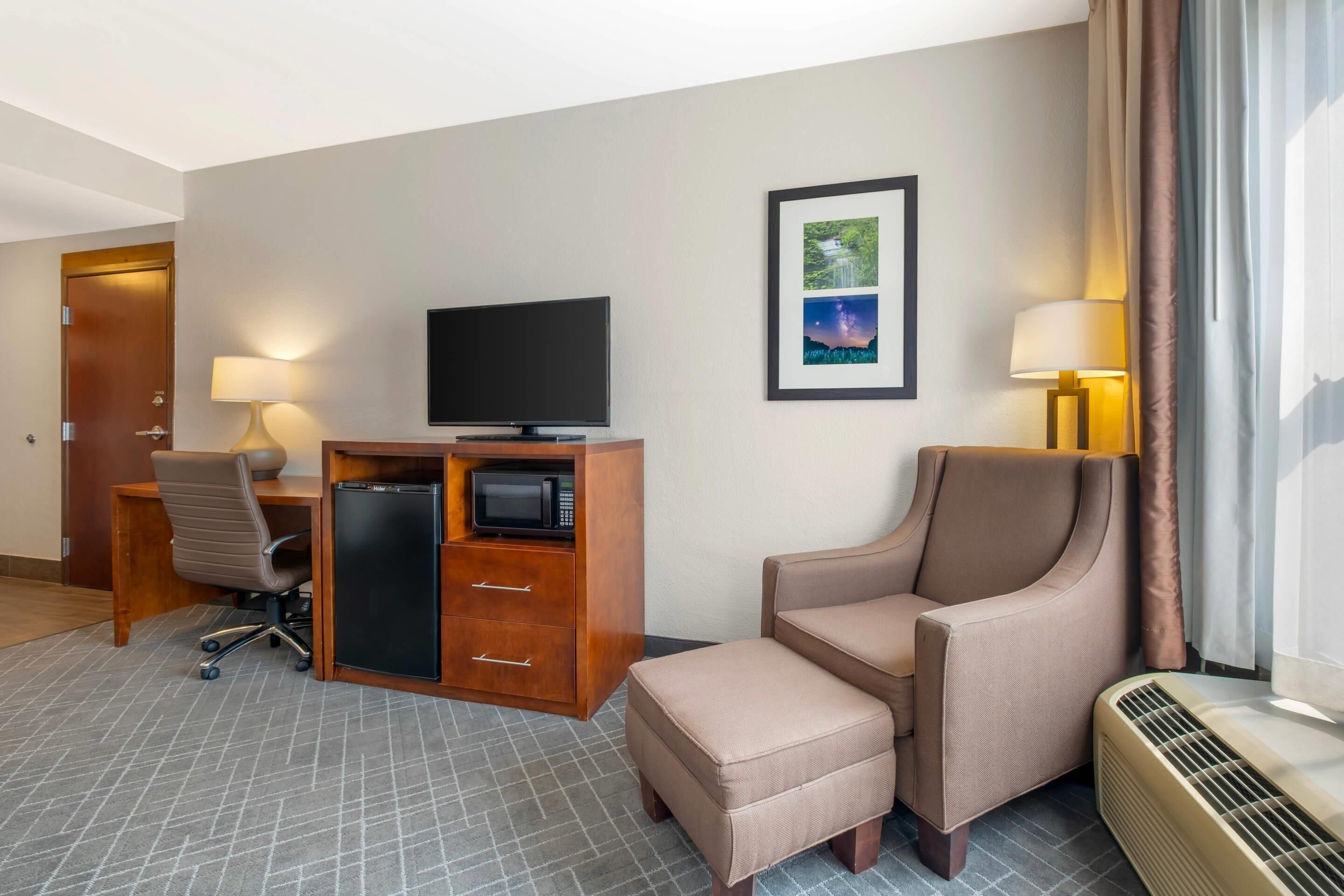 A hotel room with a desk, chair, television, microwave, and a large window with curtains.
