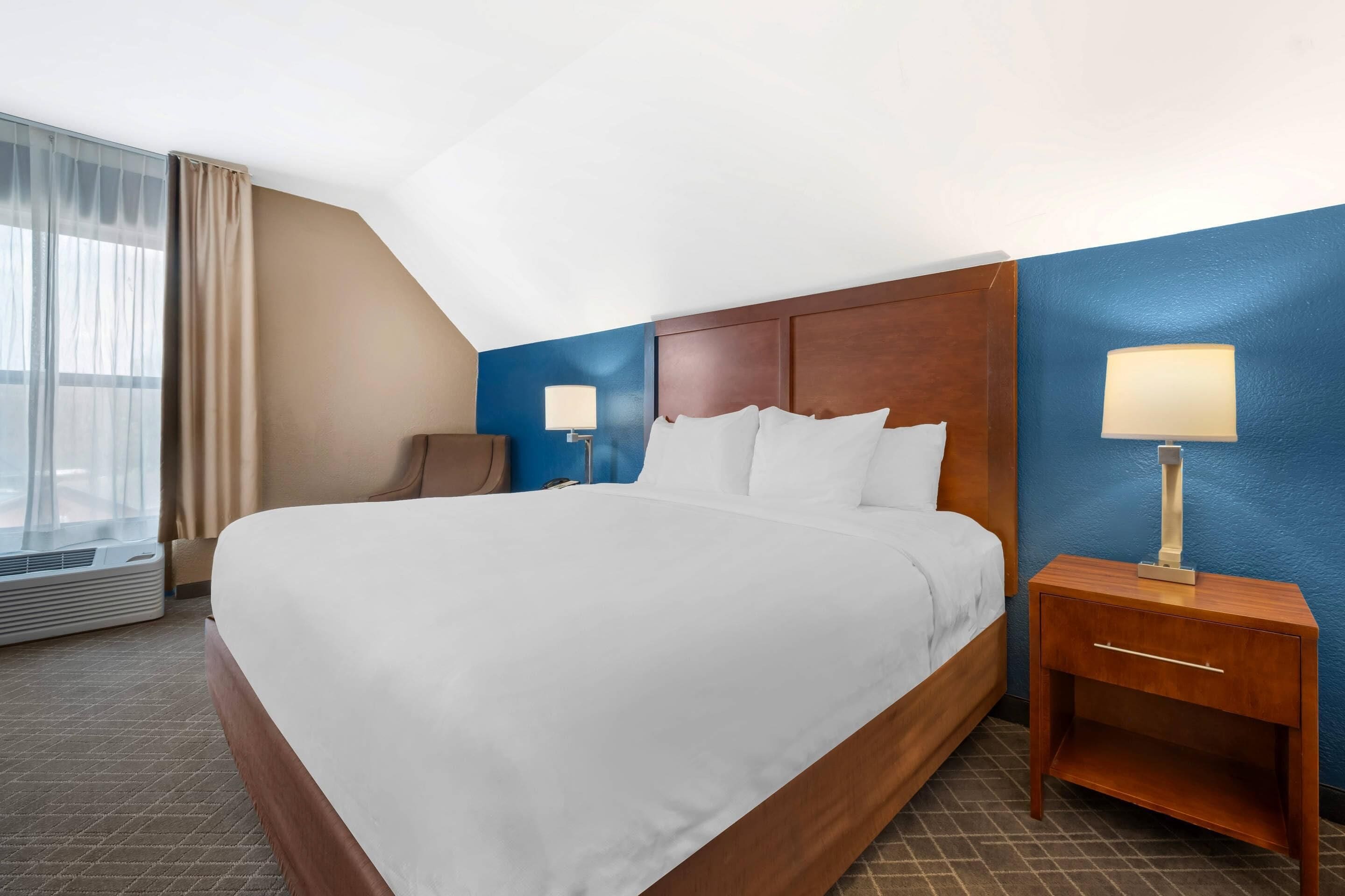 A hotel room with a large bed, two bedside tables with lamps, a window with curtains, and an air conditioning unit.