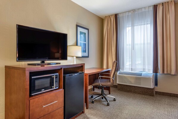 A hotel room with a TV, microwave, mini-fridge, desk, chair, air conditioner, and a window with curtains.
