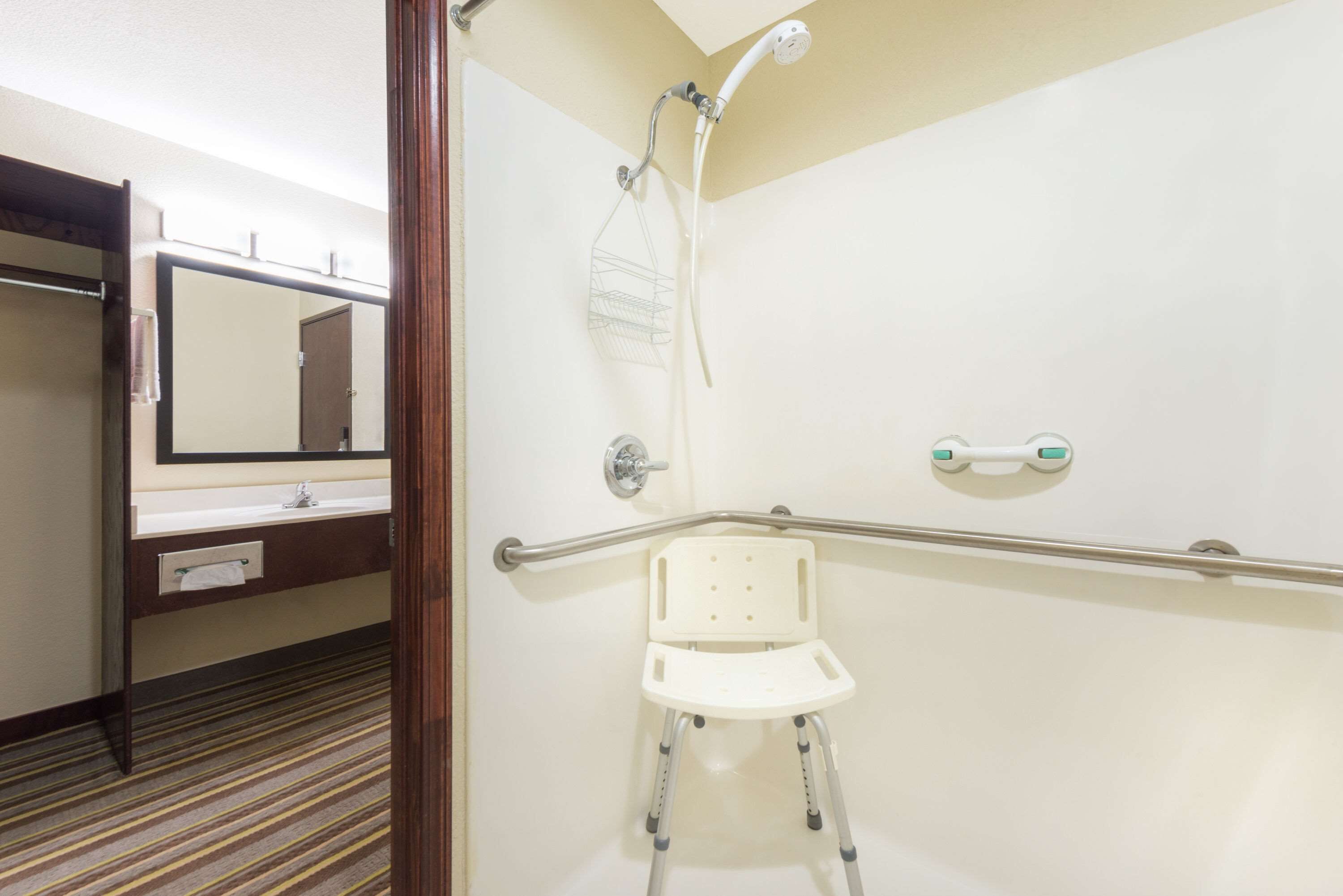 A bathroom with a shower, a handrail, a white chair, a mirror, and a sink.