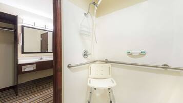 A bathroom with a shower, a handrail, a white chair, a mirror, and a sink.