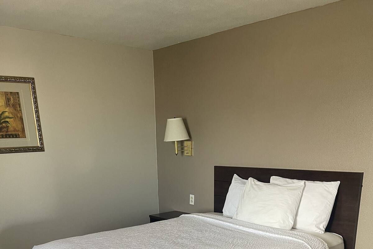 Un lit bien fait, recouvert d’une courtepointe blanche à texture, avec deux oreillers, une lampe de chevet et un tableau encadré accroché au mur.