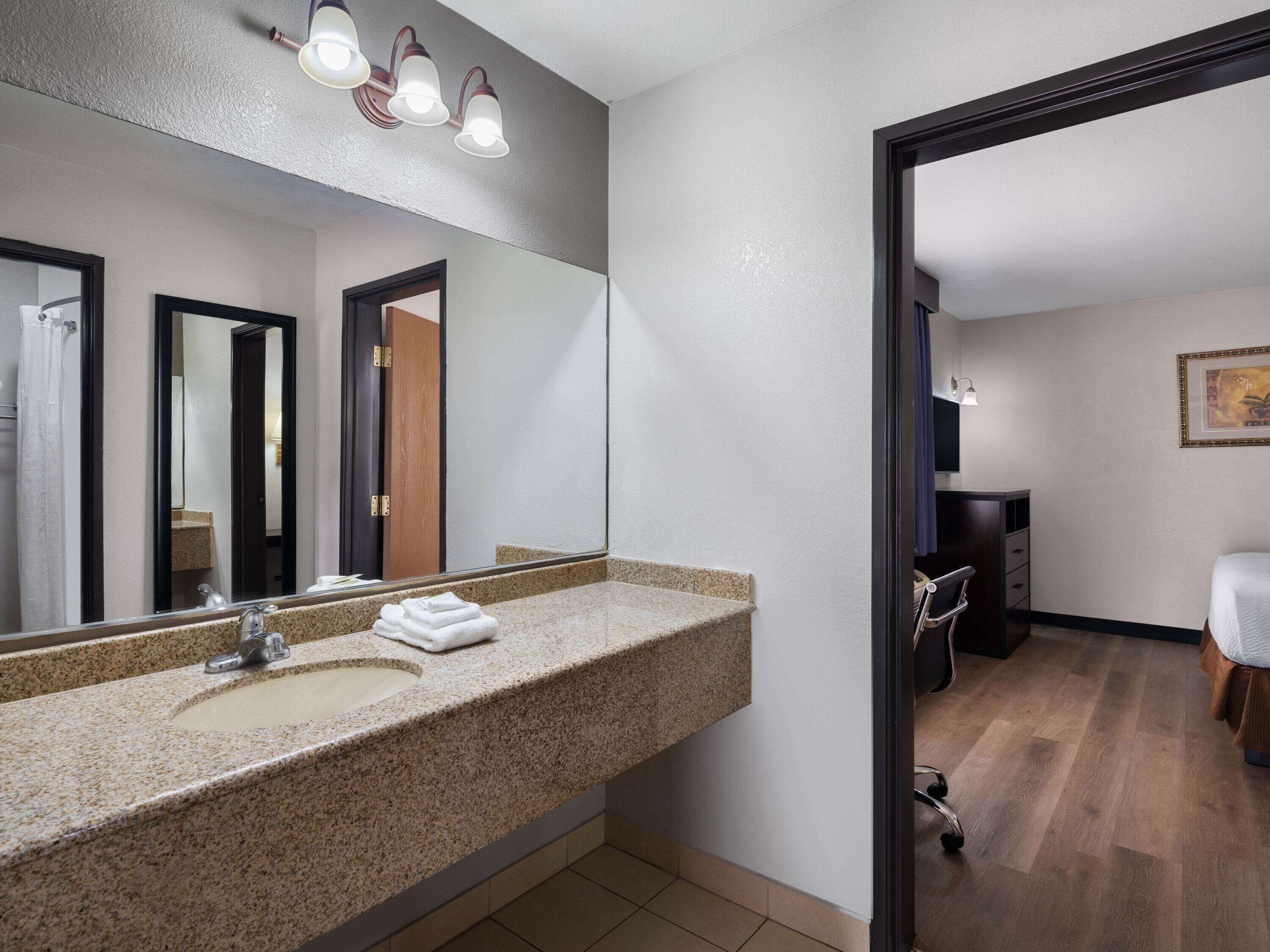 A hotel bathroom with a granite countertop sink, a large mirror, and a wooden door leading to a bedroom.