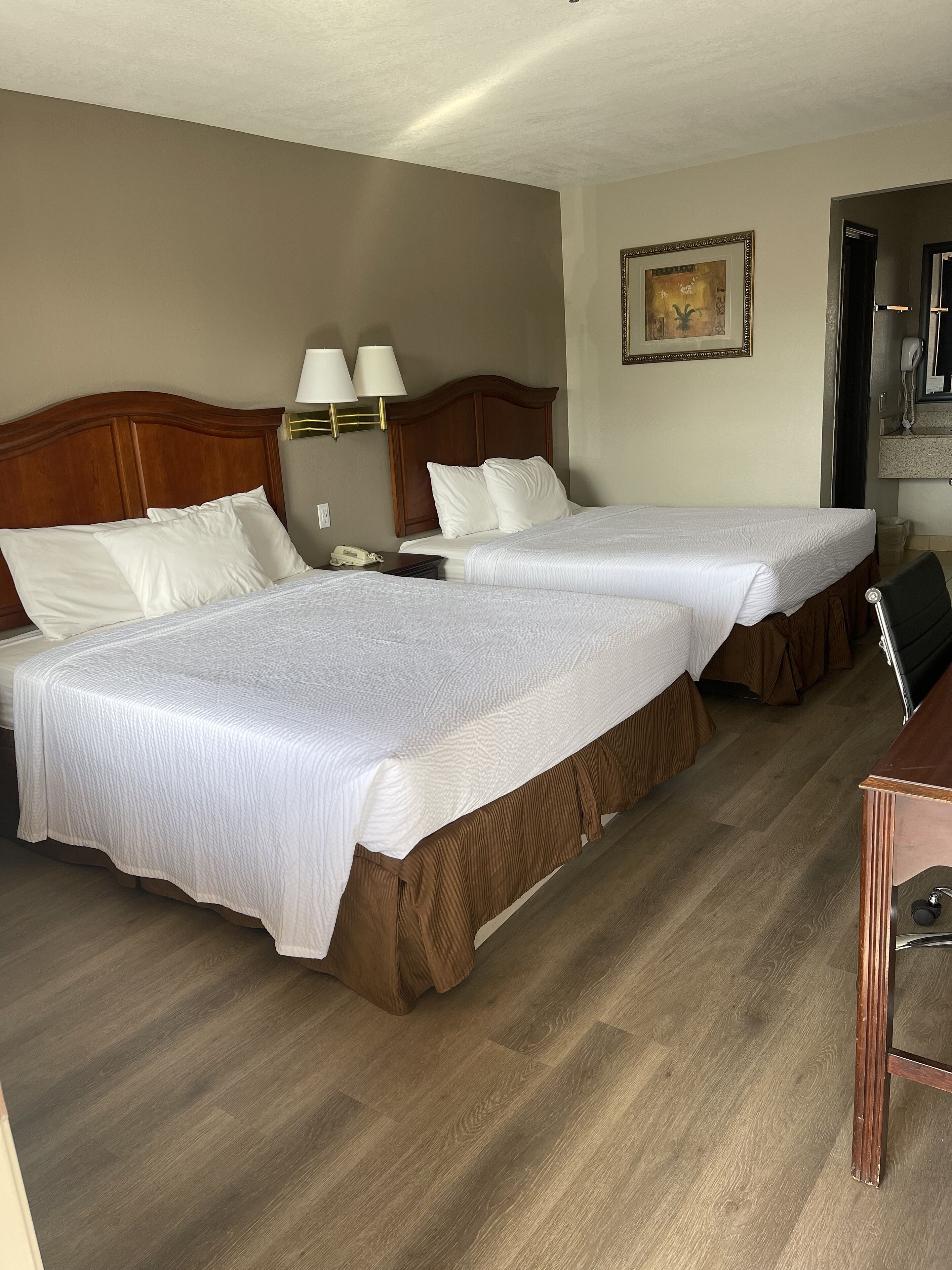A hotel room with two beds, a desk, a chair, a lamp, and a painting on the wall.