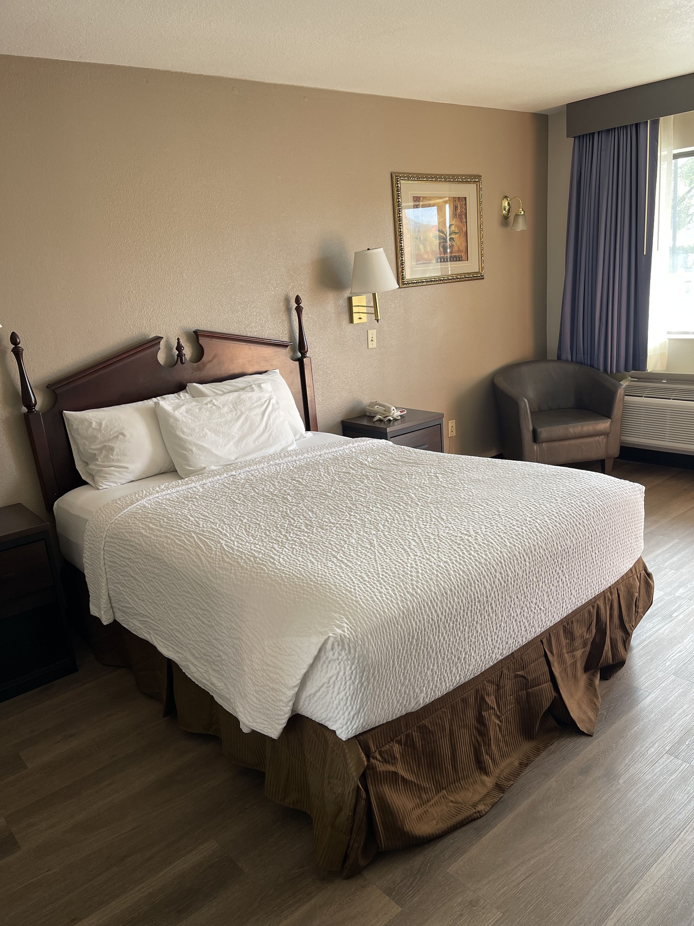 Una camera d'albergo con un letto grande, un comodino, una sedia, una lampada a parete e un quadro incorniciato.