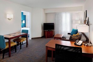 A hotel room with a dining table, chairs, a television, a sofa, a lamp, and a telephone.
