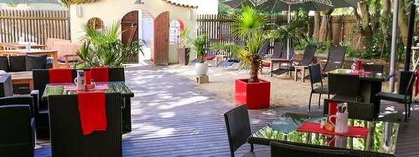 Un área de patio exterior con mesas y sillas, algunas con detalles en rojo, y plantas en macetas.