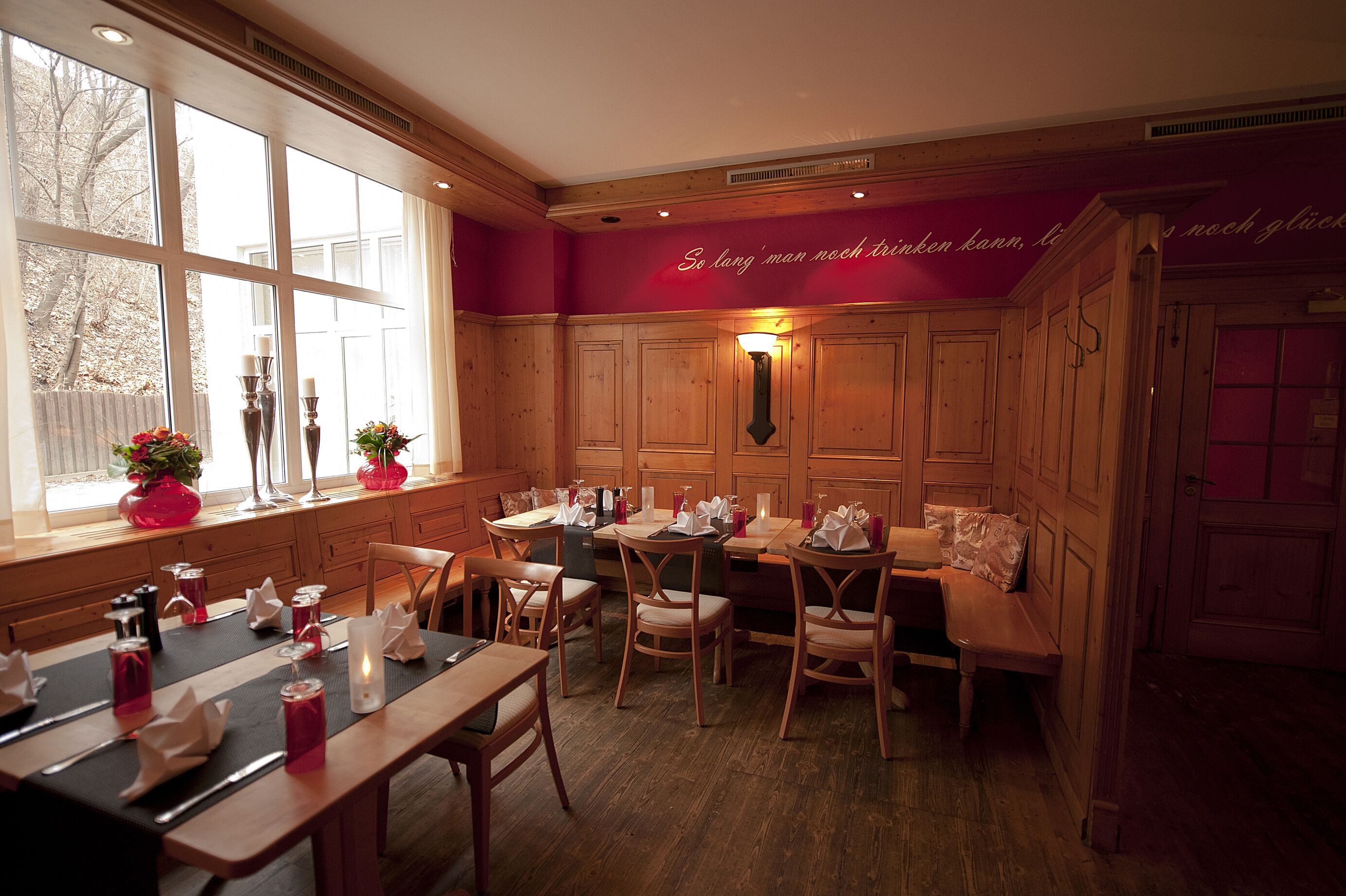 Un restaurante con paneles de madera, ventanas amplias y una pared roja con texto. Las mesas están dispuestas con velas y flores.