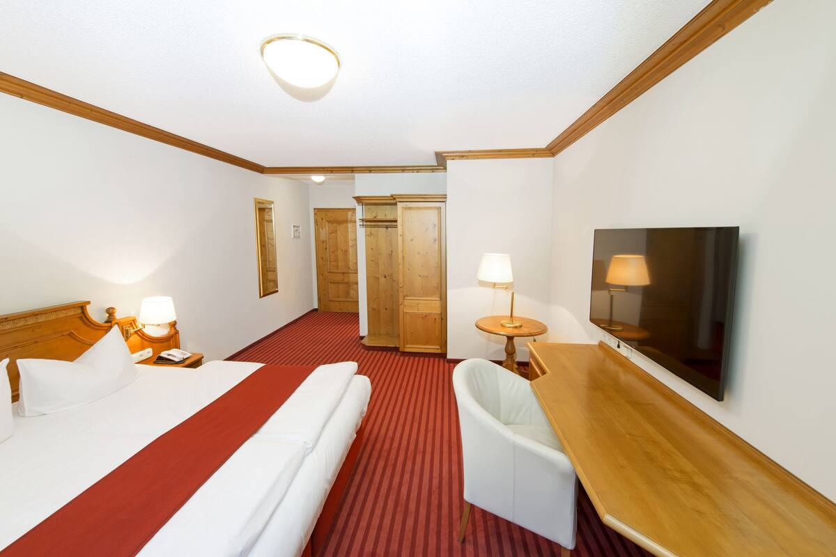 Hotellihuone, jossa on sänky, yöpöydät, työpöytä, tuoli, televisio ja ovi, joka johtaa toiseen huoneeseen.