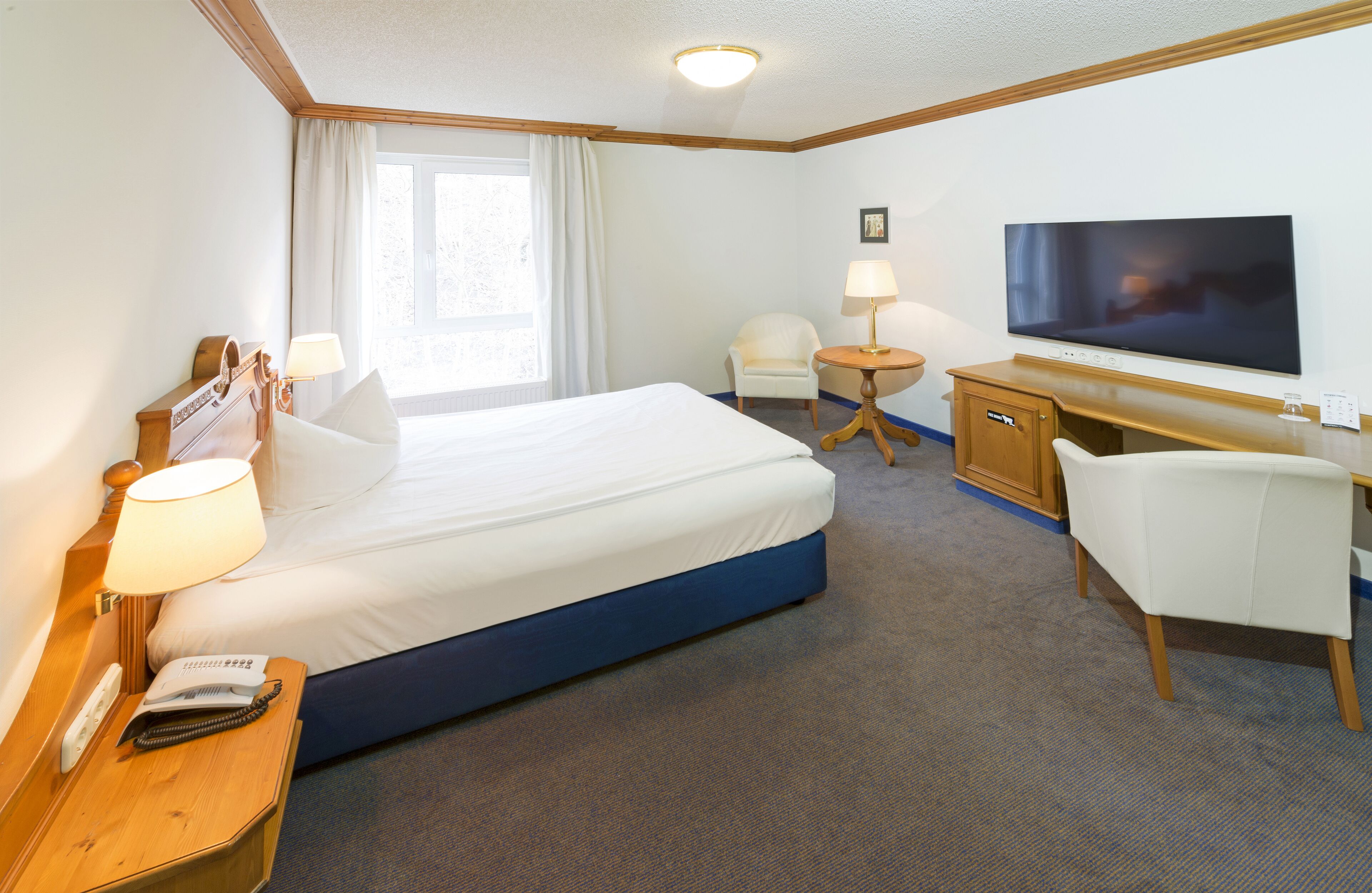 Ein Hotelzimmer mit Bett, Nachttisch, Lampe, Schreibtisch, Stuhl, Fernseher und einem fenster mit Vorhängen.