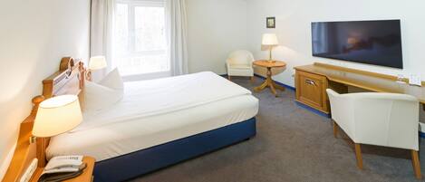 Ein Hotelzimmer mit Bett, Nachttisch, Lampe, Schreibtisch, Stuhl, Fernseher und einem fenster mit Vorhängen.
