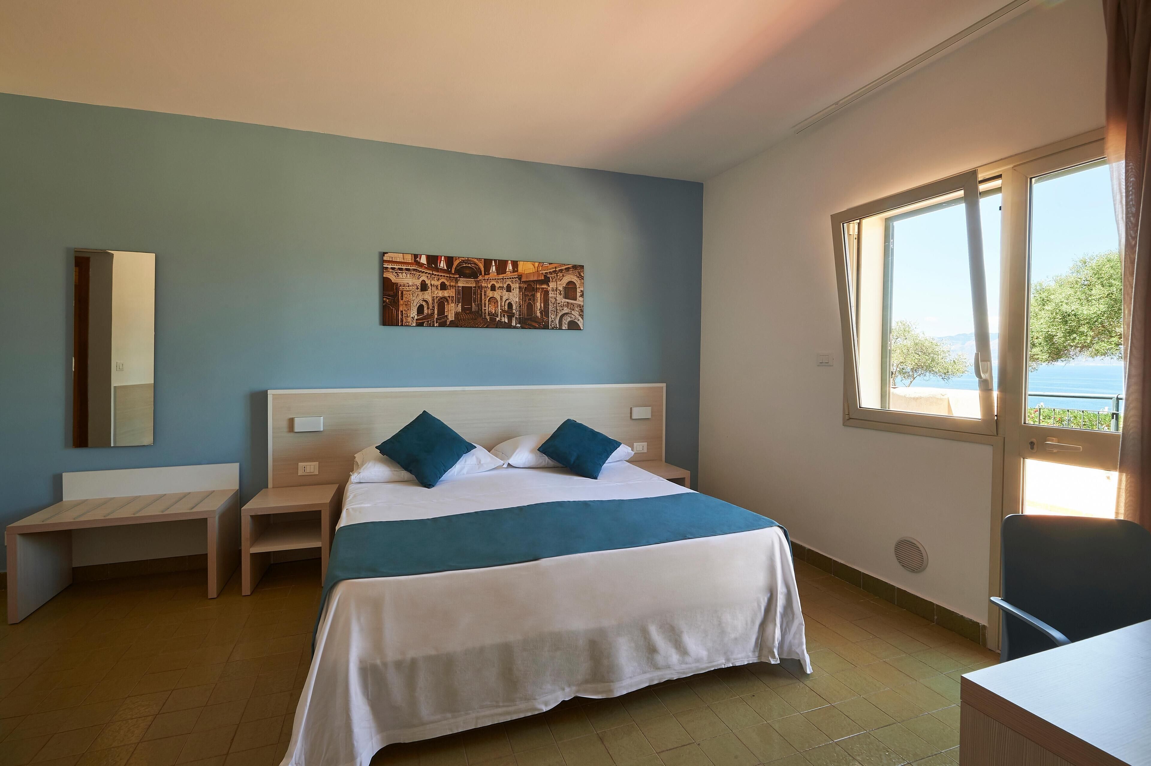 Una habitación de hotel con cama, escritorio, silla, ventana con vistas y un cuadro en la pared.