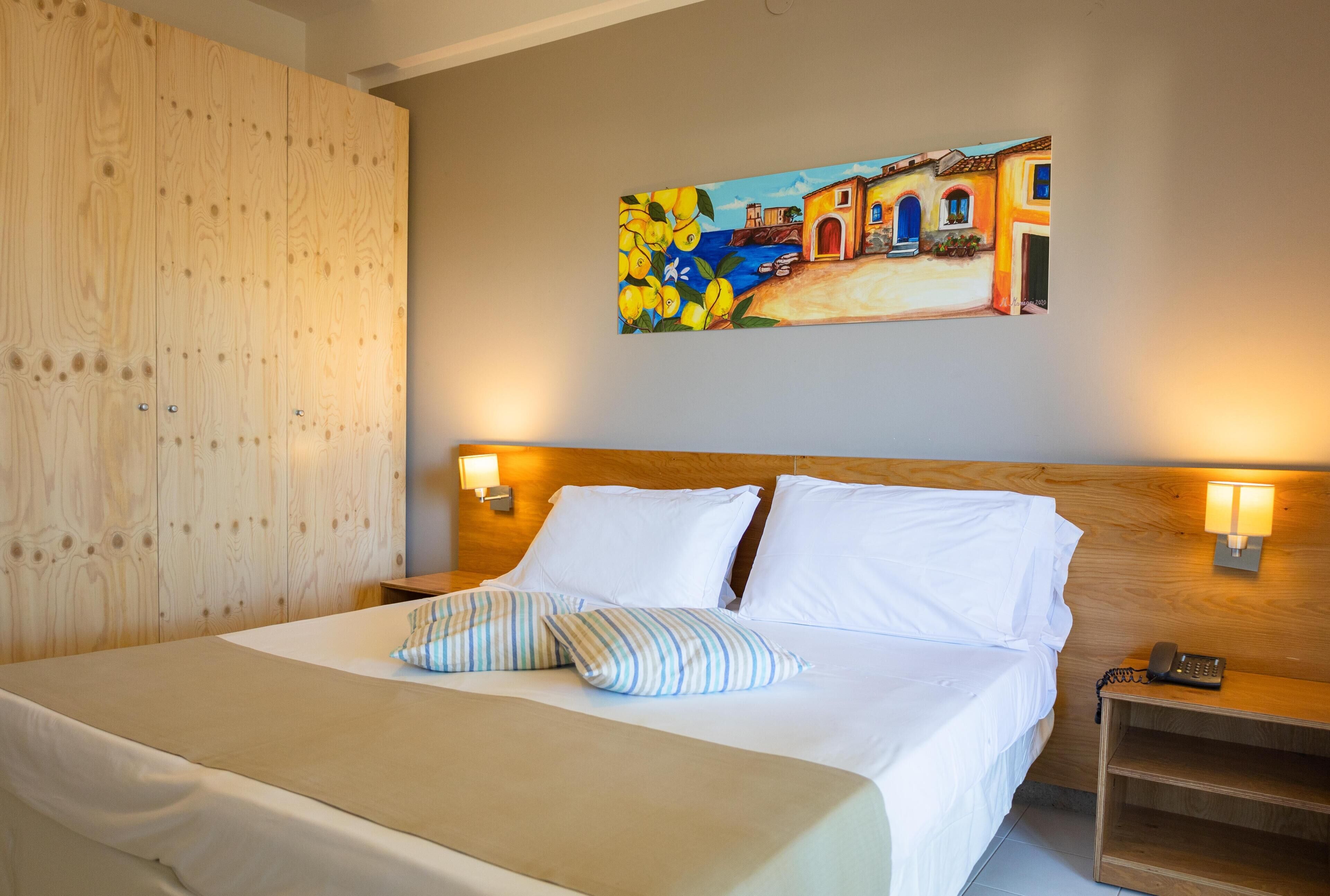 Habitación de hotel con cabecera de madera, cama con sábanas blancas, almohadas a rayas y un cuadro con una escena costera en la pared.