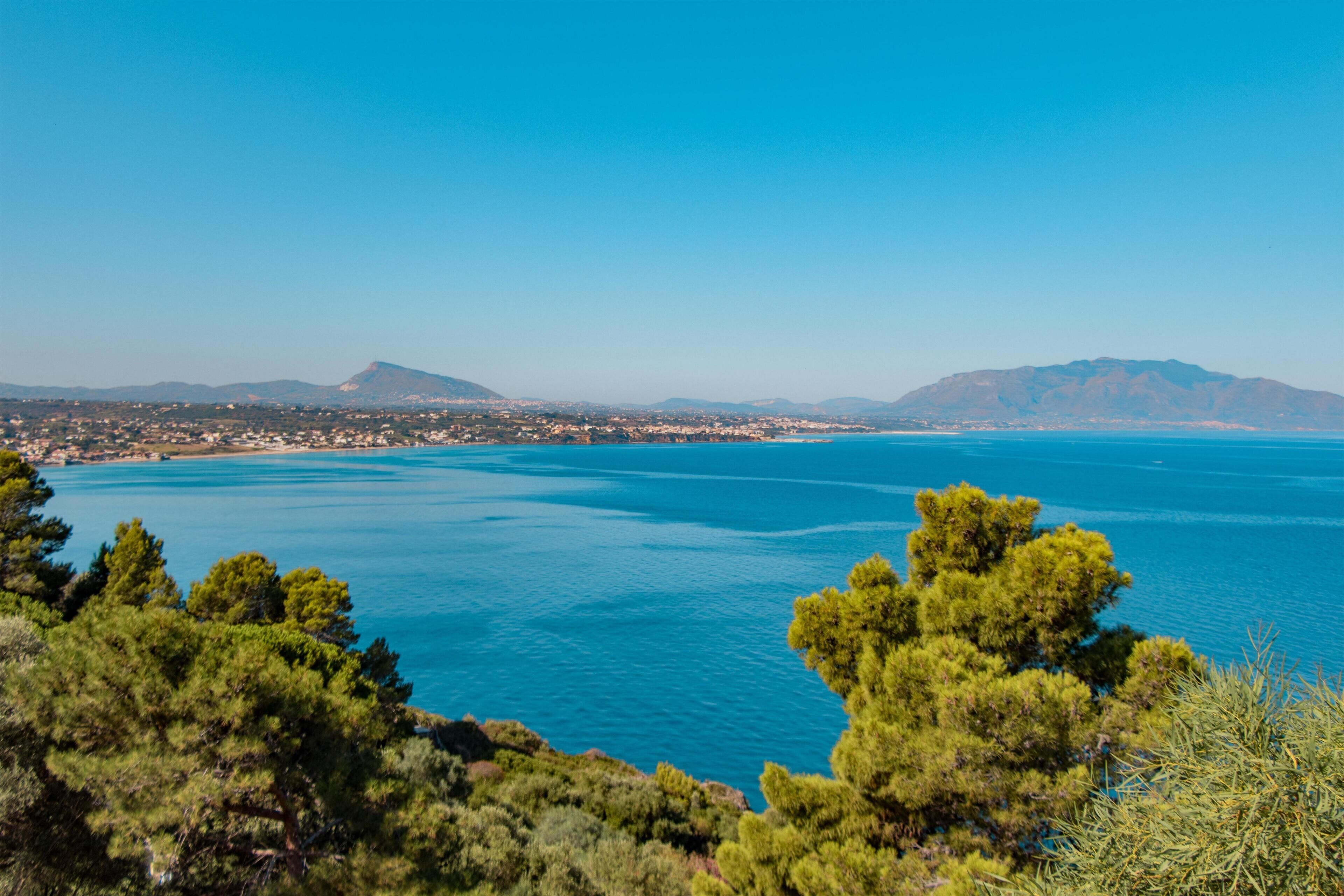 Paesaggio costiero con vista limpida sul mare, montagne in lontananza e vegetazione fitta in primo piano.