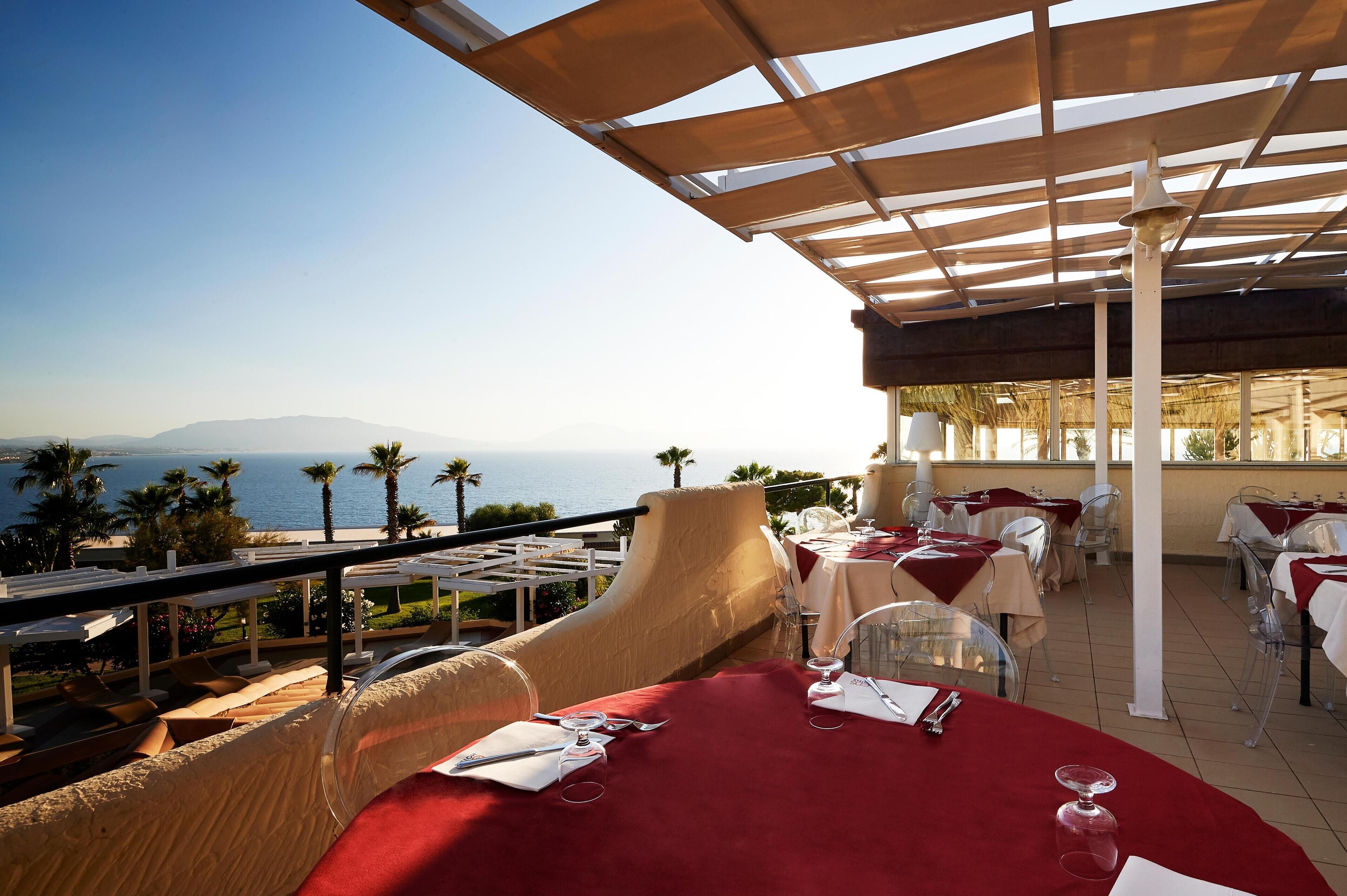 A terrace with tables set for dining, overlooking a coastal view with palm trees and mountains.