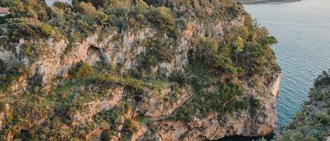 Un acantilado costero con la entrada natural a una cueva, rodeado de vegetación densa y vistas a una extensión de agua.