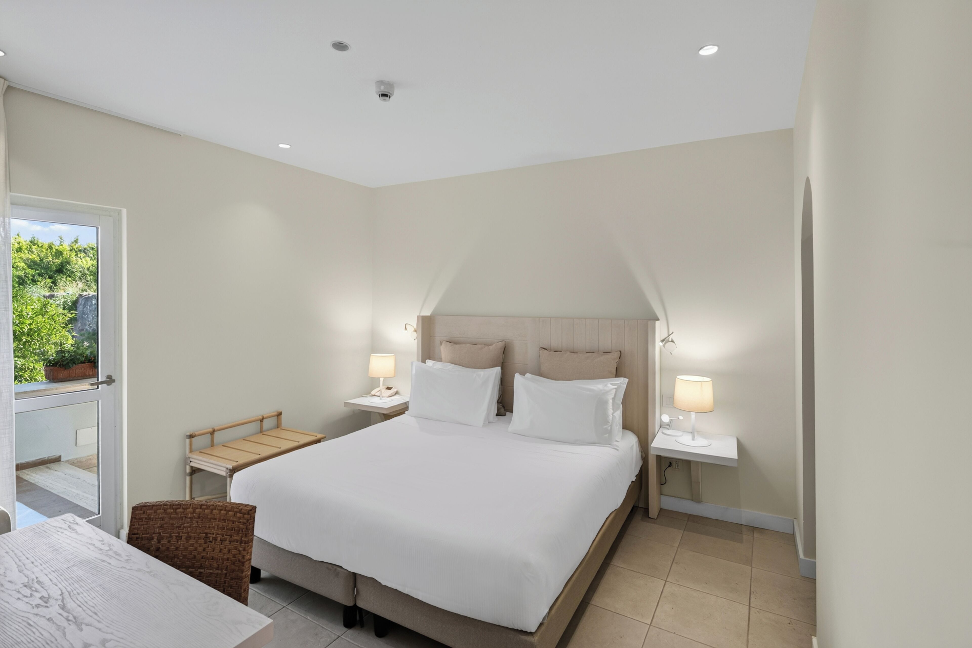 Un dormitorio moderno con una cama grande, mesitas de noche con lámparas, un pequeño rincón de descanso y vistas a zonas verdes.