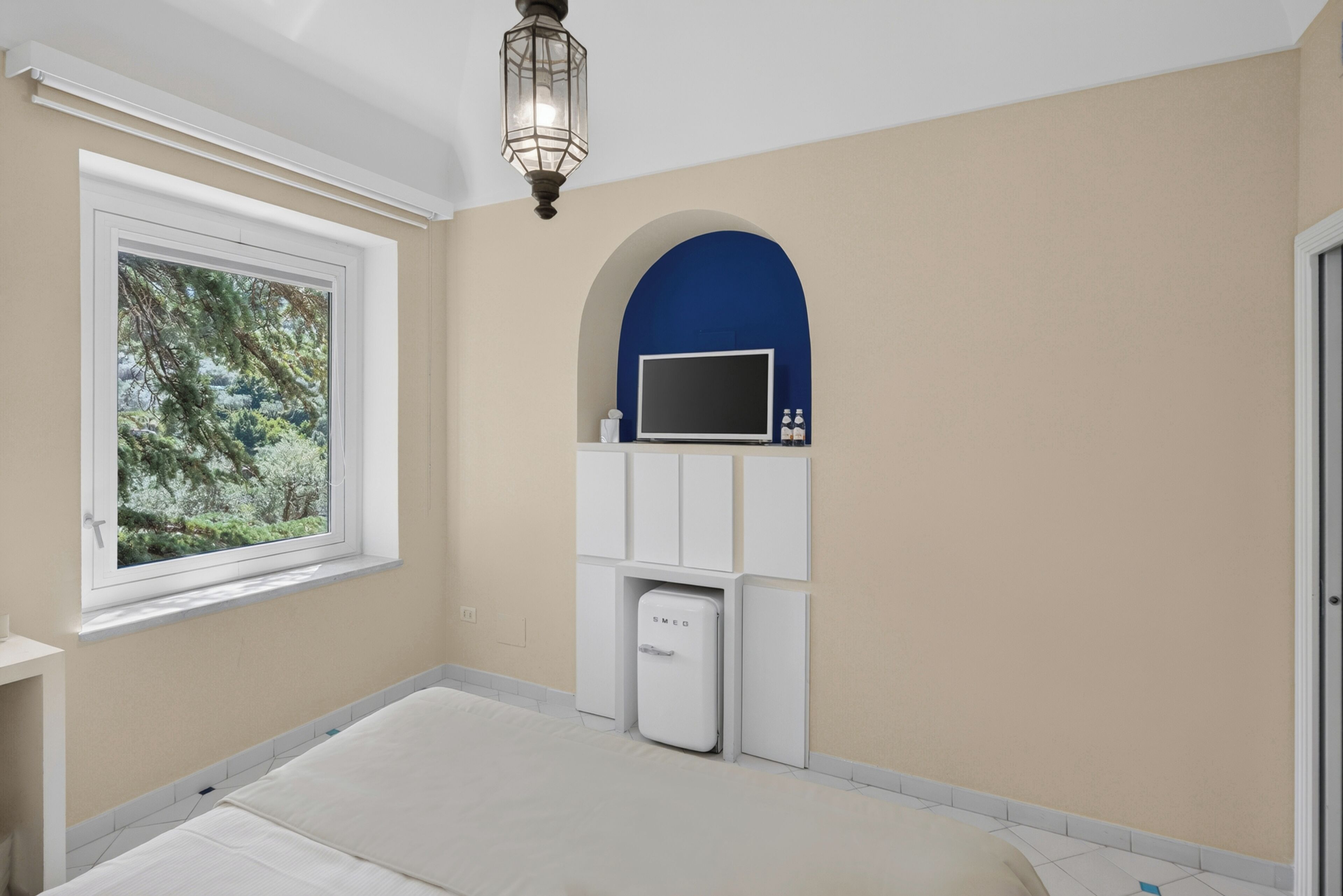 A bedroom with a bed, a TV mounted on a blue arch, a white cabinet, and a small fridge.